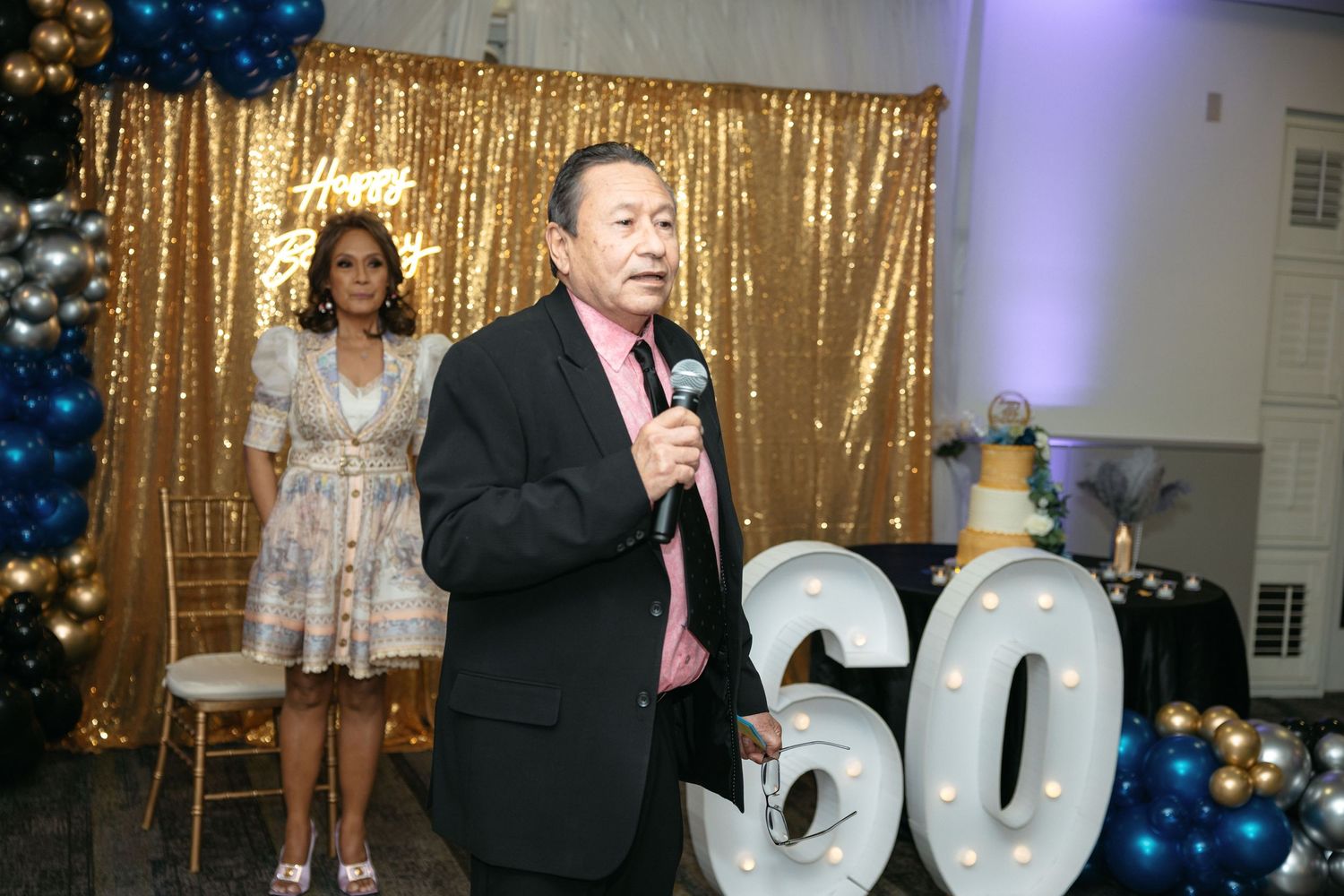 A speaker addresses guests at a 60th birthday celebration with gold sequin backdrop and decorative numbers.