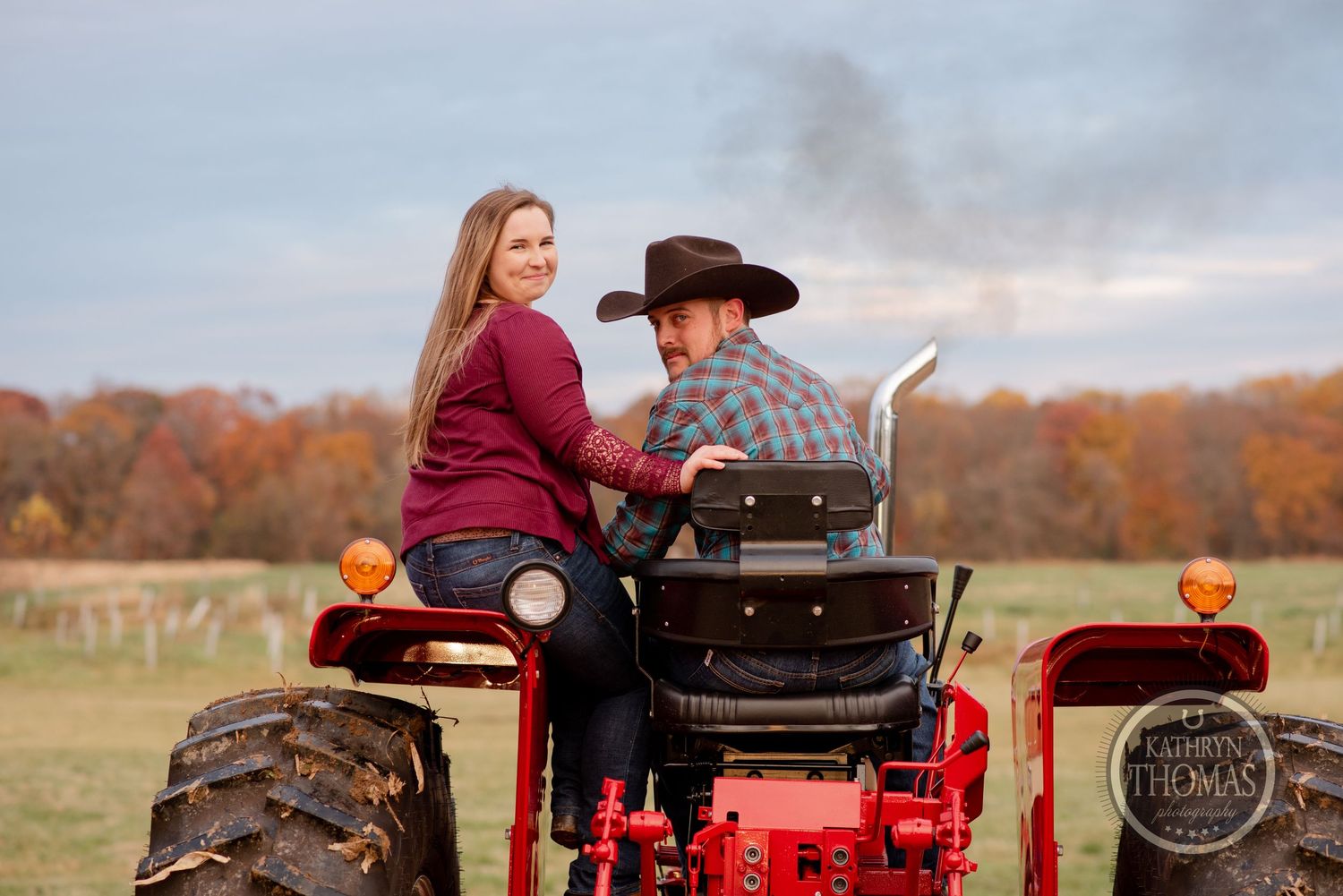 A couple sits together on a red tractor during an autumn photoshoot with fall foliage in the background.
