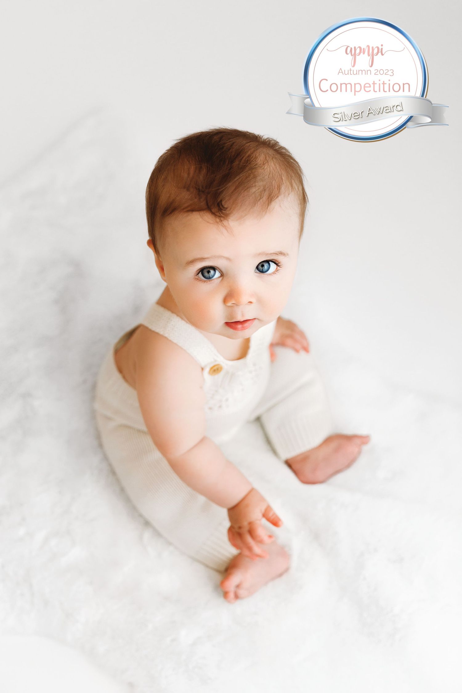 Sitting baby boy, wearing white overalls and looking up with big bright blue eyes. Silver Award-winning image