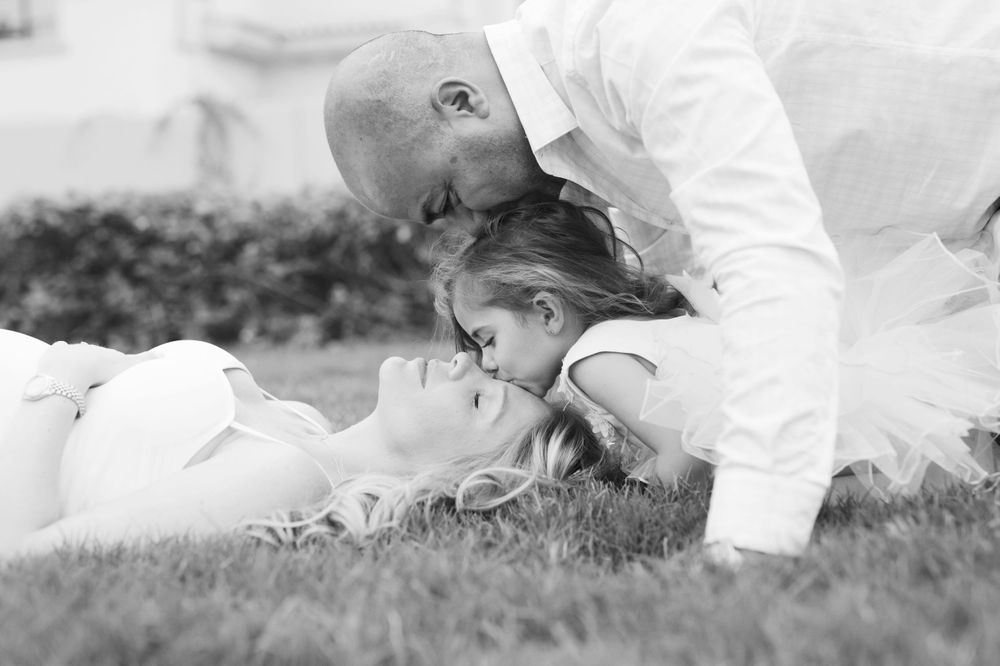 Mom, Dad and little girl posing on the grass, each placing a kiss on the others head.