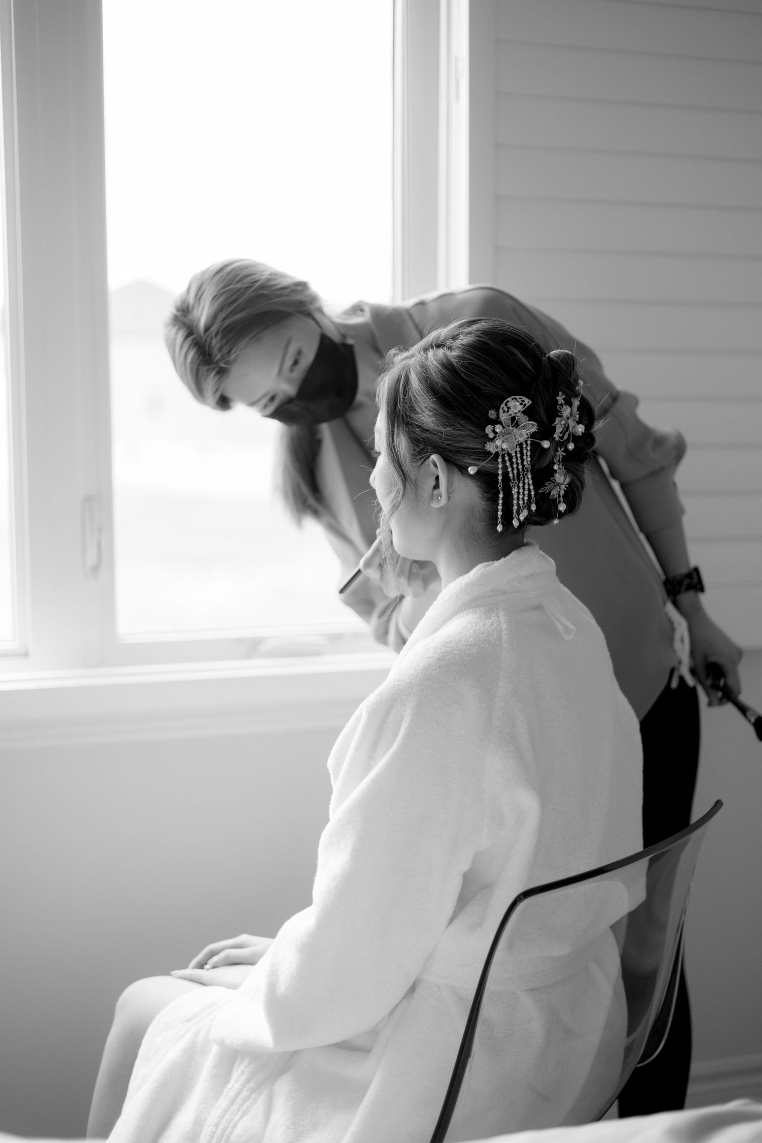 Black and white image of stylist working on intricate bridal hairstyle near window.