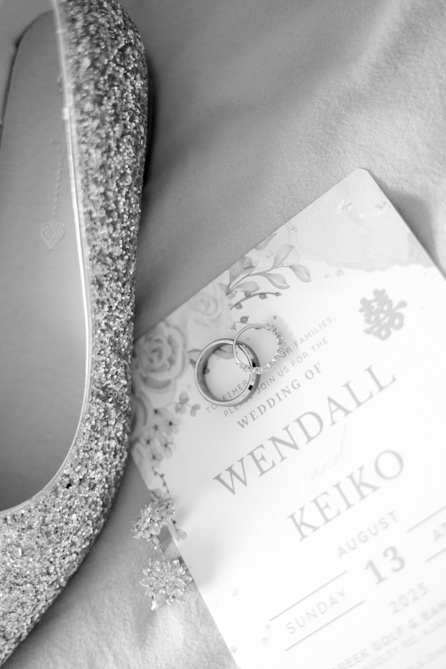 Wedding invitation and sparkling silver high heel shoe with wedding rings arranged on white surface.