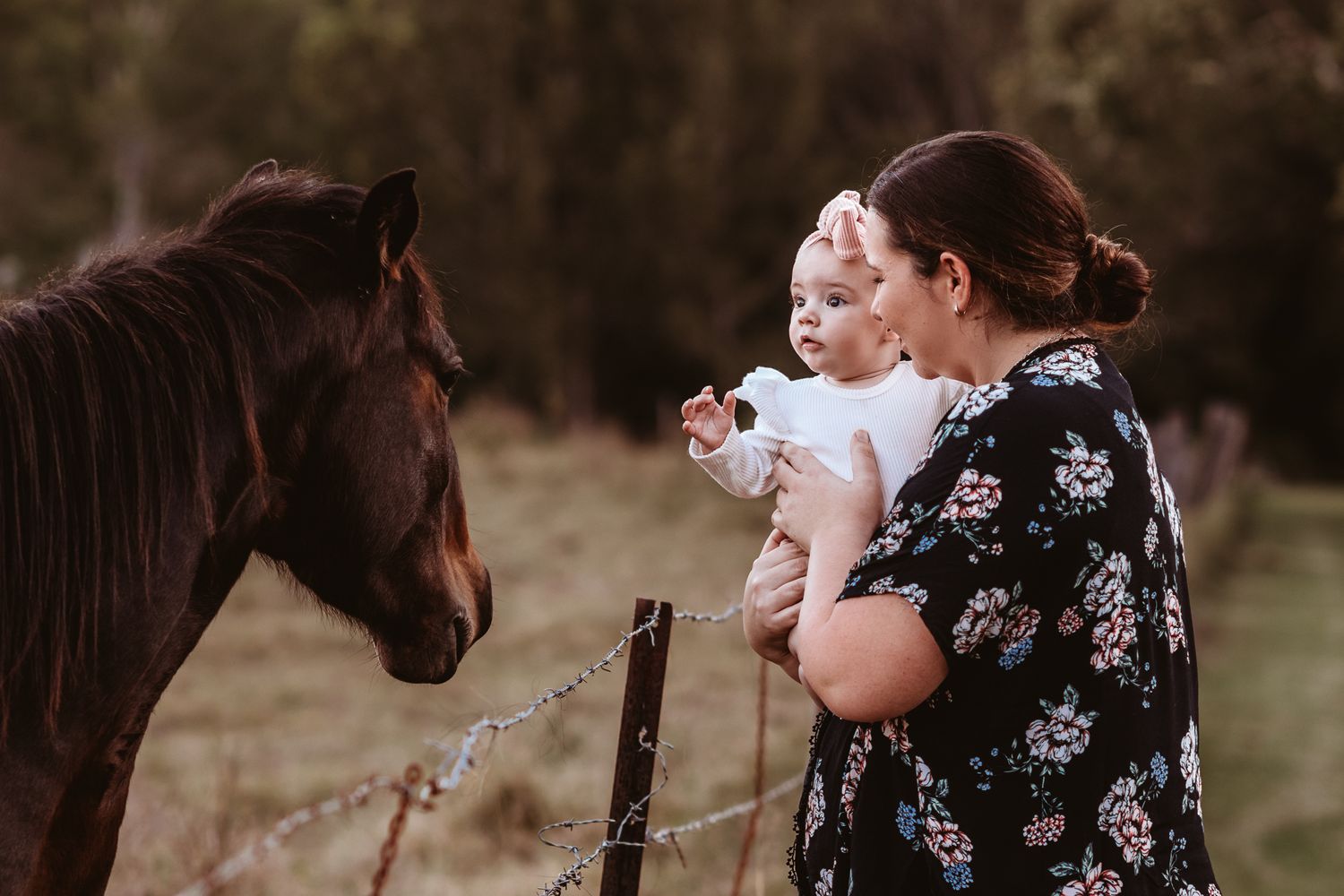 A brown horse meets a child and someone wearing a floral dress in a pastoral outdoor setting.