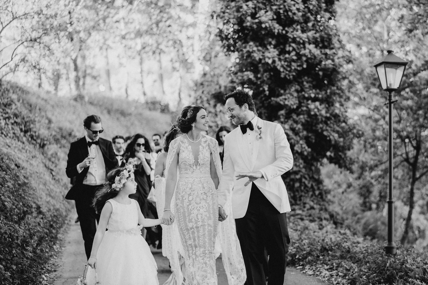 A black and white photo of wedding guests walking along a tree-lined path.