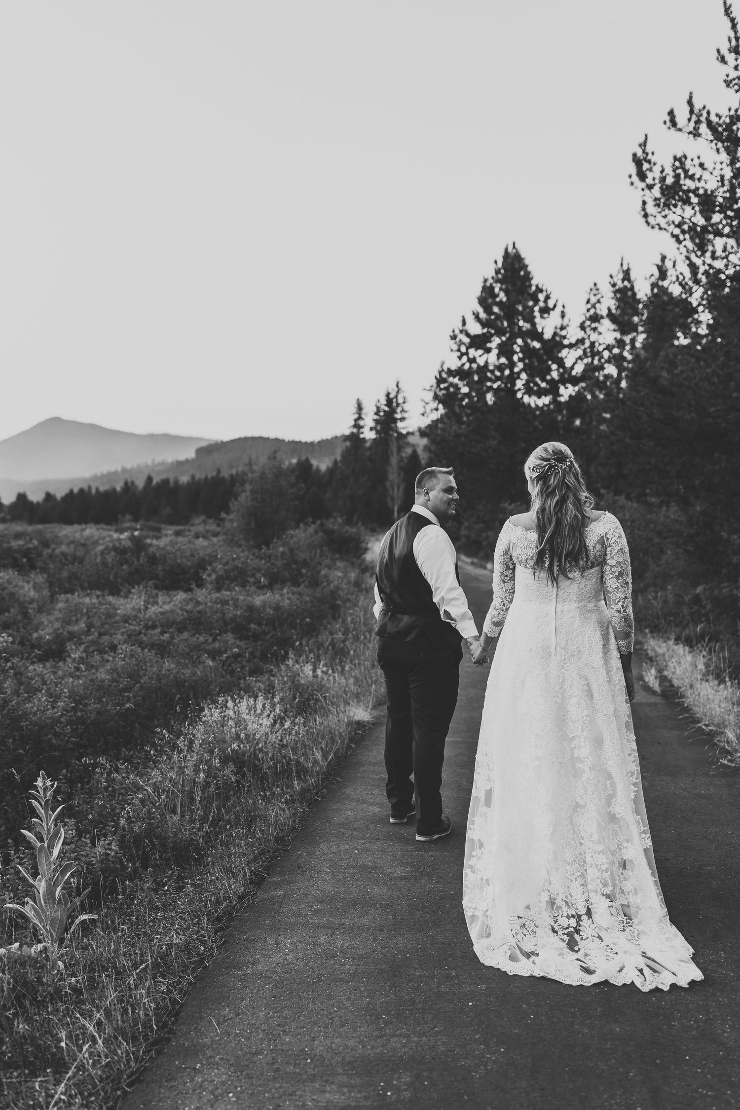 A couple in wedding attire holds hands while walking along a mountain trail at dusk.