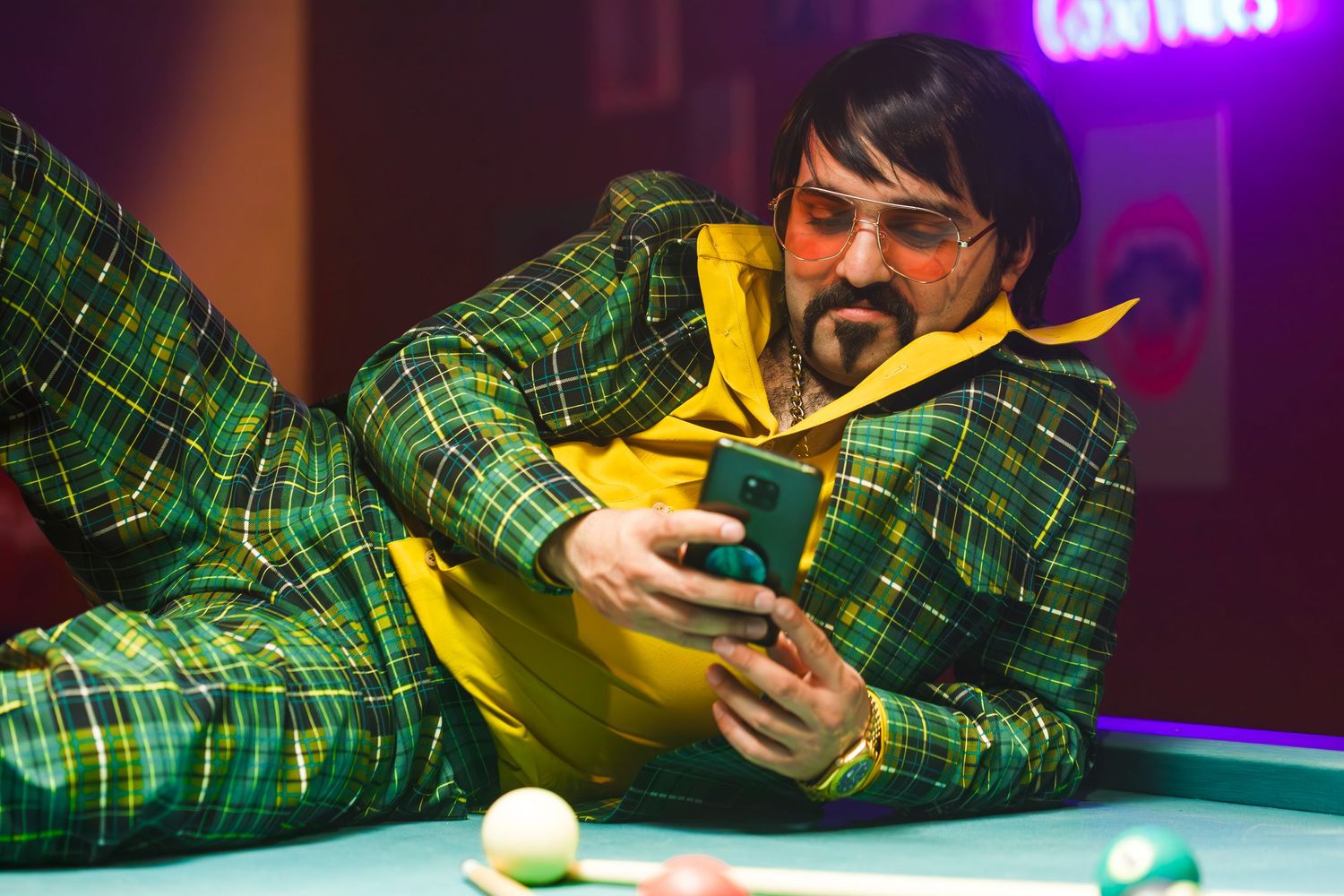 Person in green plaid suit lying on pool table with yellow accent in purple-lit room.