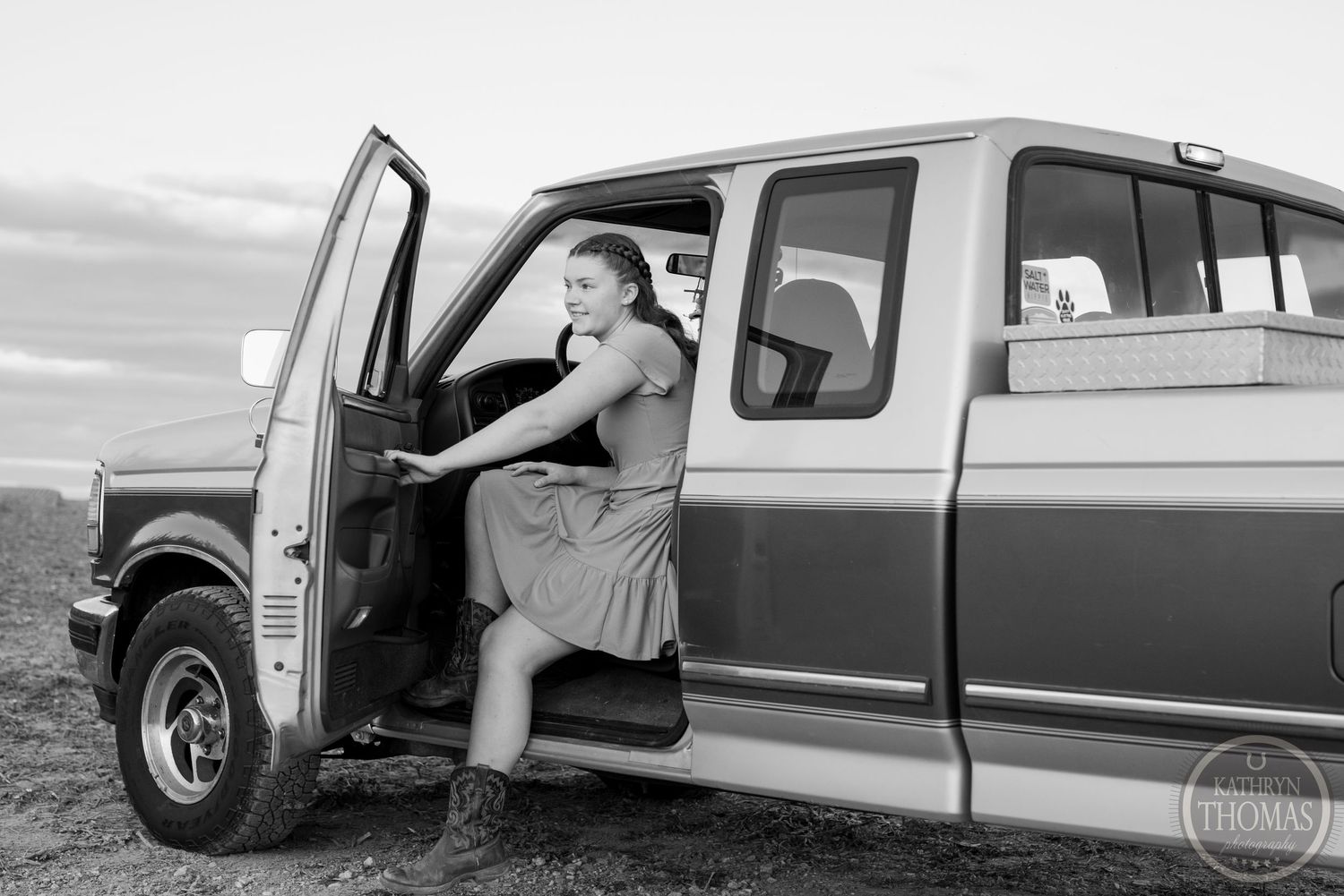 Black and white photo of someone sitting in a vintage pickup truck with the door open.