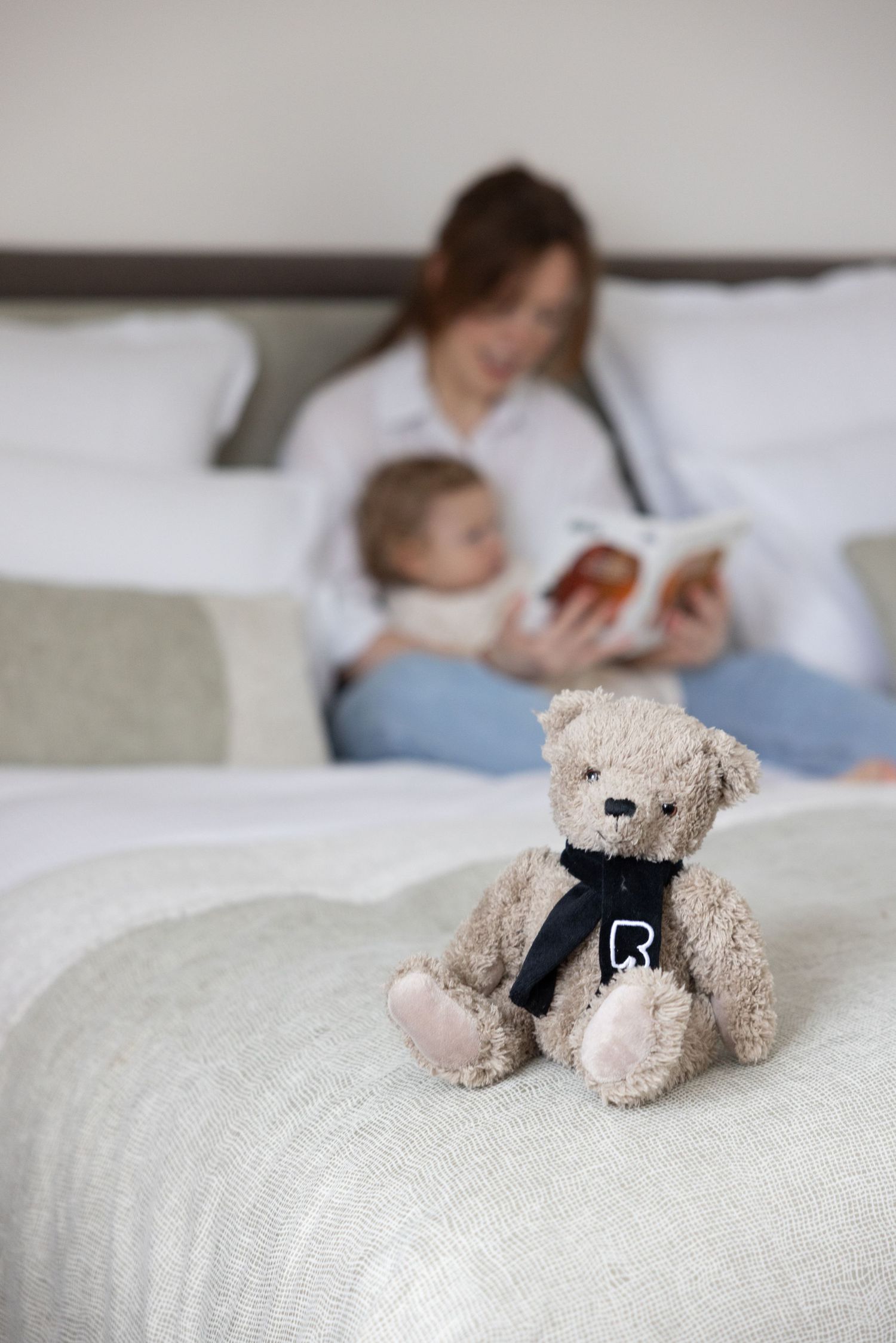 Plush teddy bear wearing black scarf sits on white hotel bed linens.