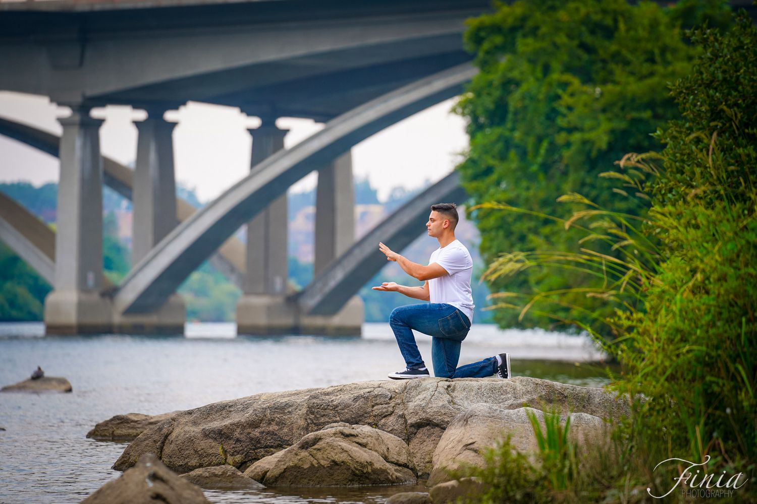 Person in a blue shirt kneels while gazing at a scenic river view with a large bridge in the background.