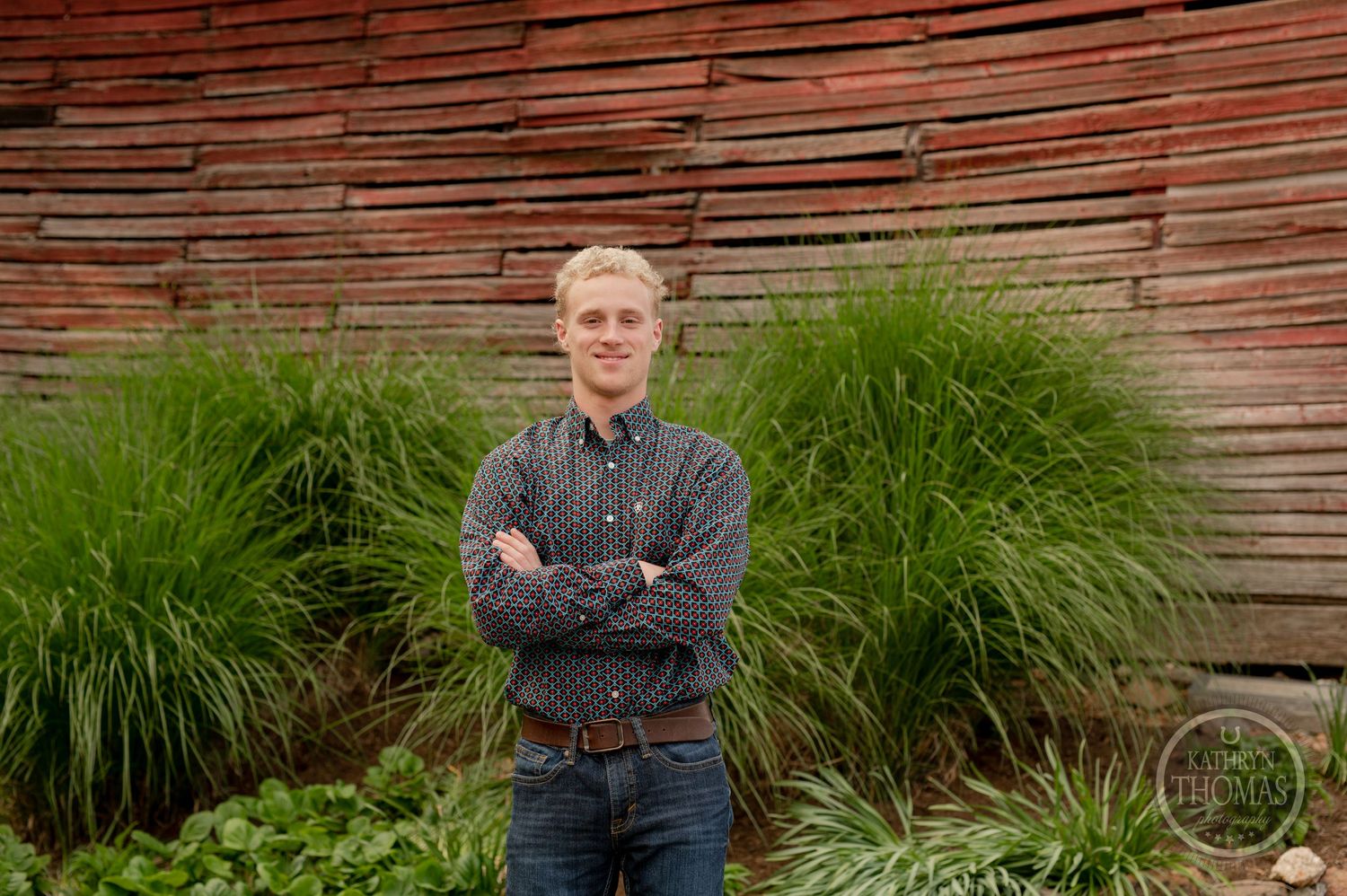 Portrait shot of person in casual attire standing against weathered red barn with ornamental grasses.