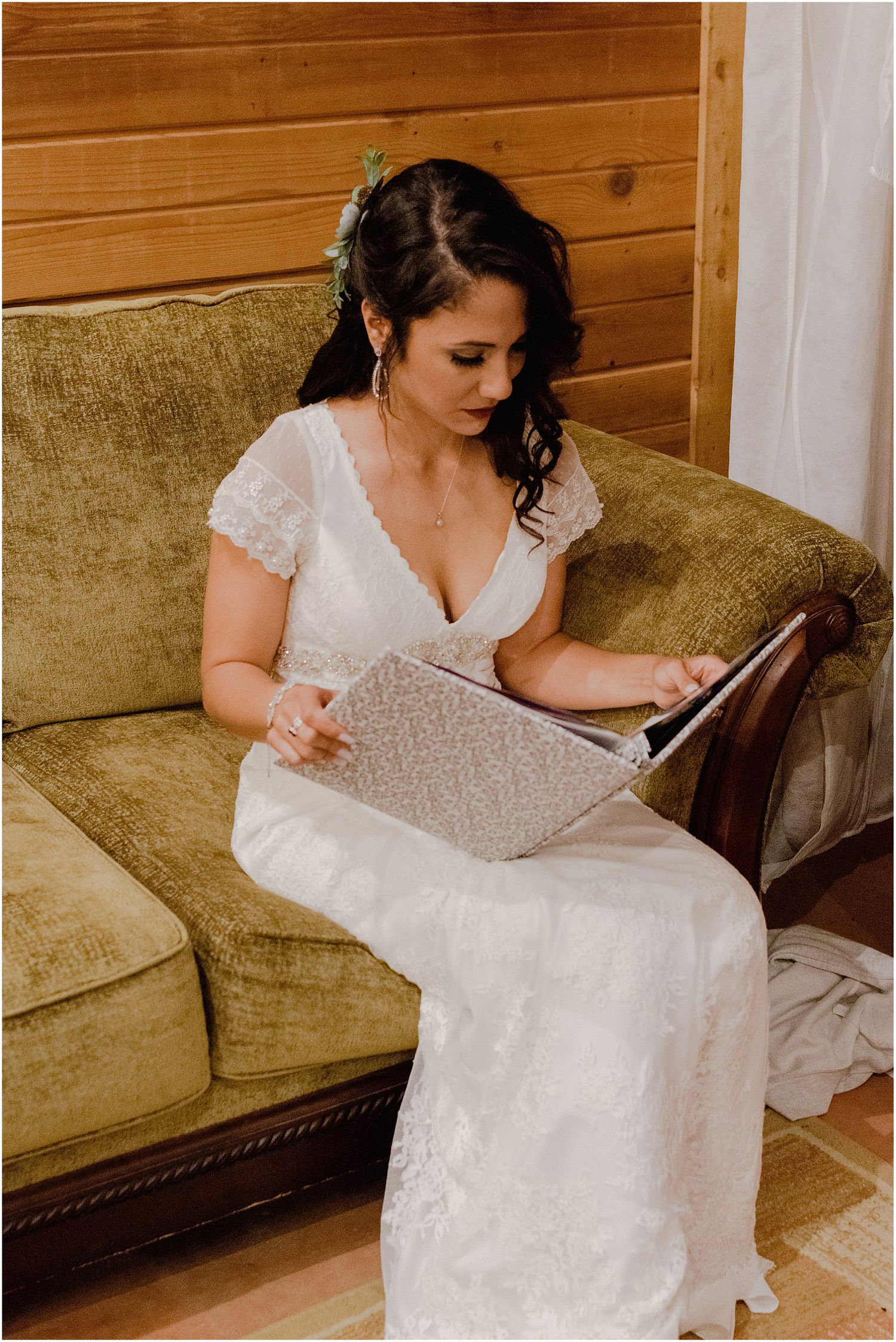 A bride in a white lace dress sits on a tan couch while looking through an album.