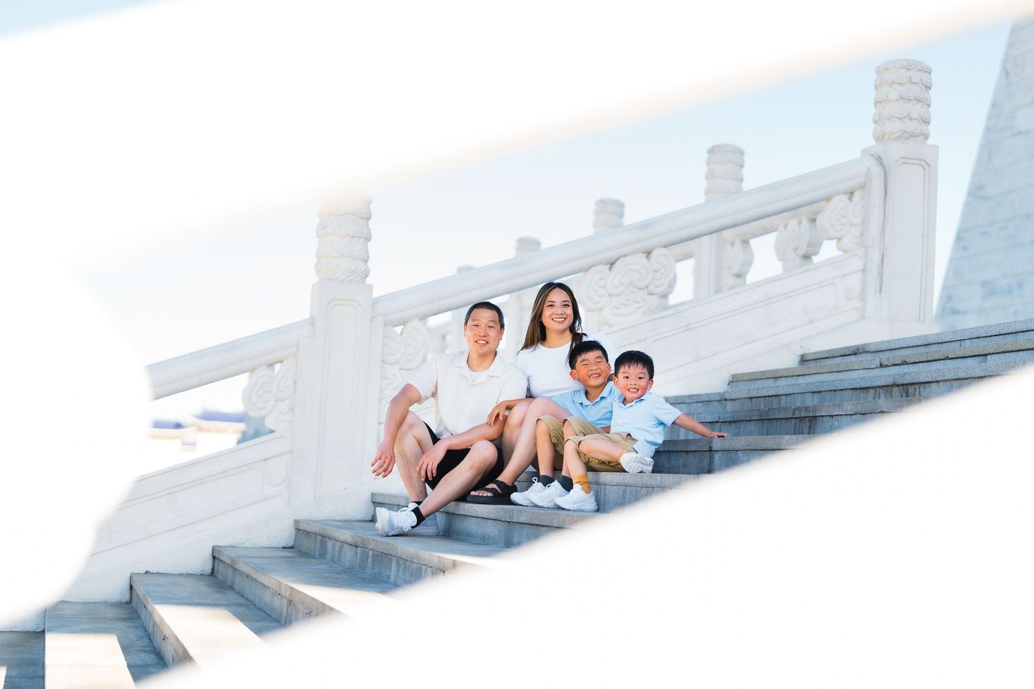 A family sits together on white stone steps outdoors on a bright sunny day.