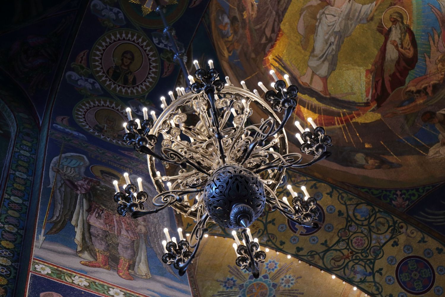 Ornate chandelier with multiple tiers of lights hanging from an orthodox church ceiling with painted religious murals.