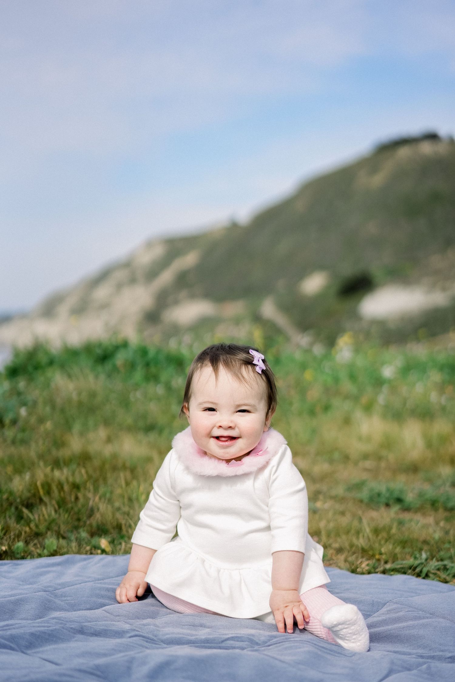 Baby in white dress sitting on blue blanket against coastal landscape.
