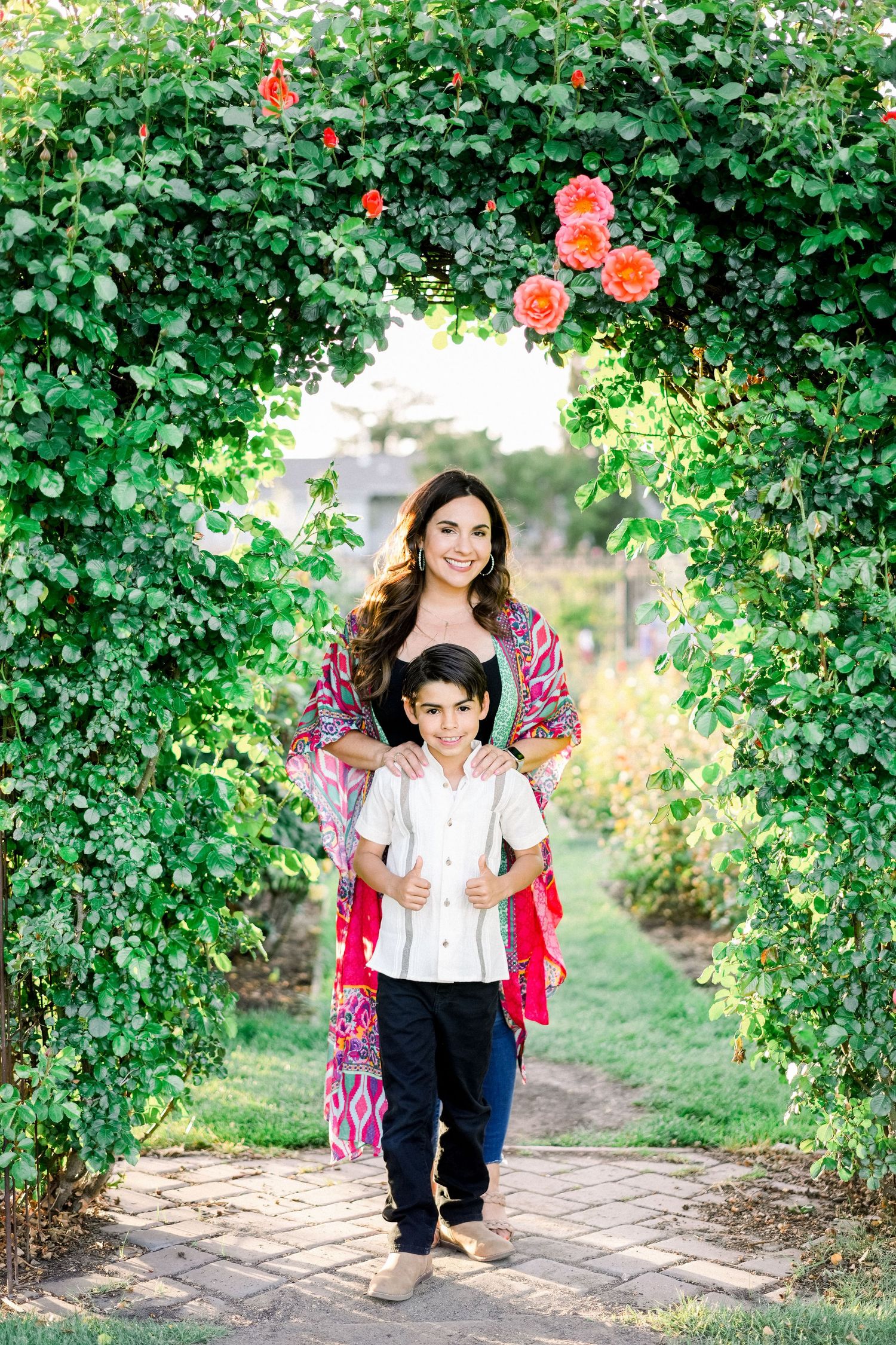 People posing under archway of flowering red roses and green vines in garden path.