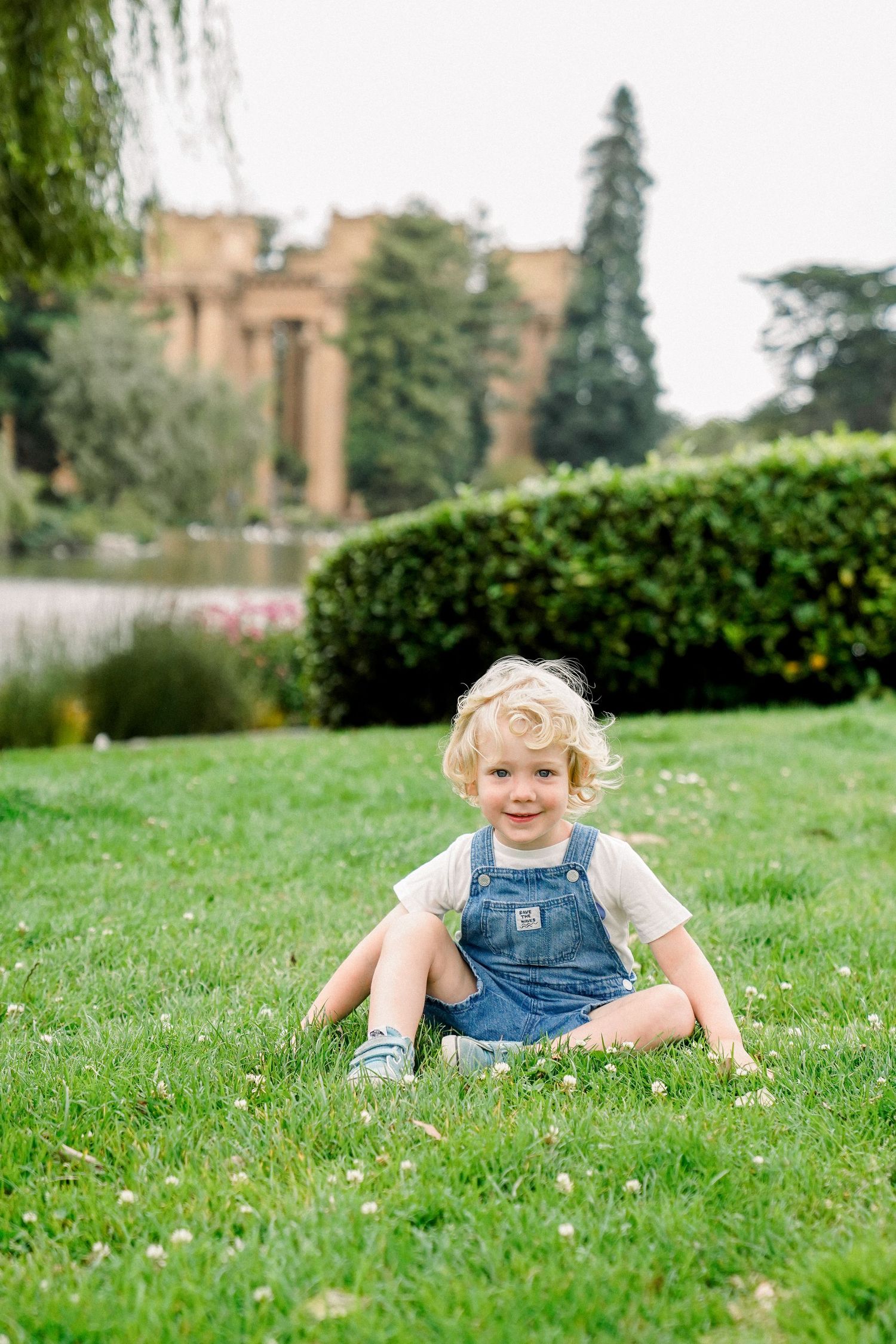 Child in denim overalls sits on grass with historic palace building in background.