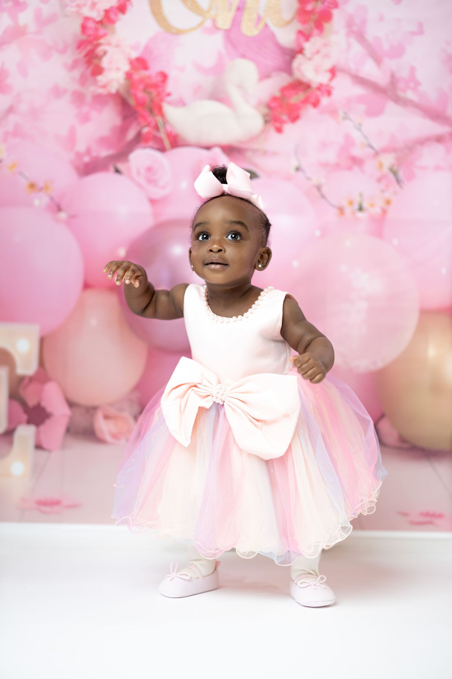 Baby in pink tulle dress celebrating first birthday with balloon backdrop.