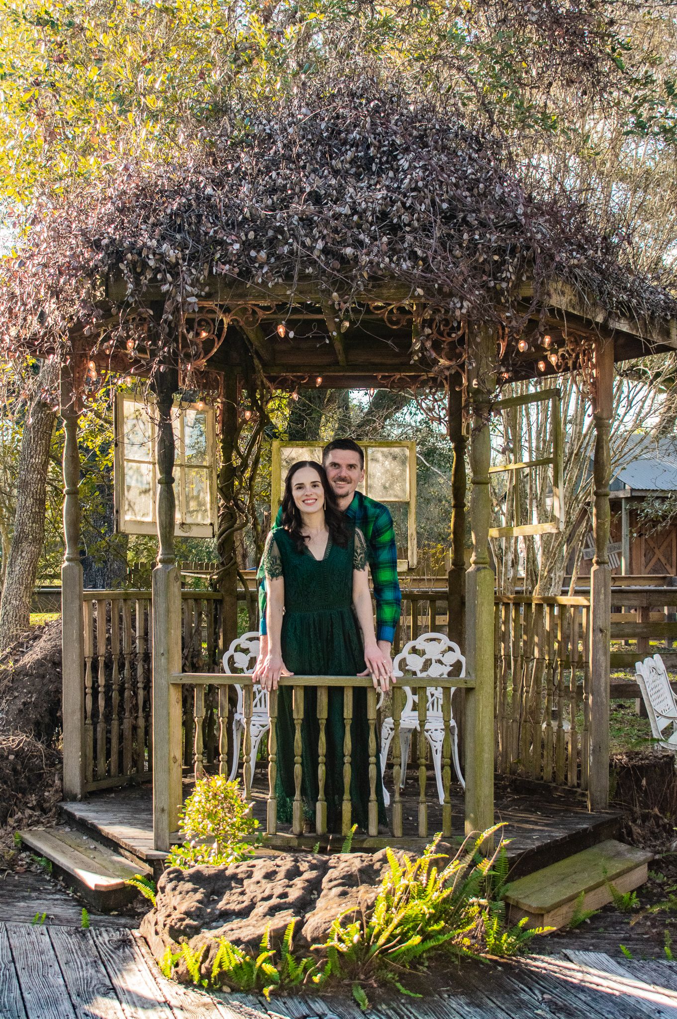 A couple stands together in a rustic wooden gazebo covered in vines.