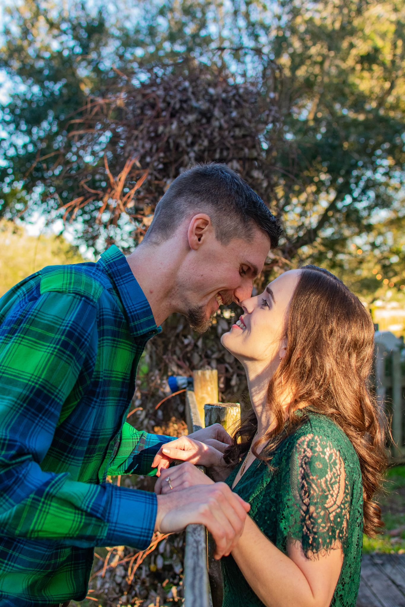Romantic couple in green attire share a playful moment by rustic fence.