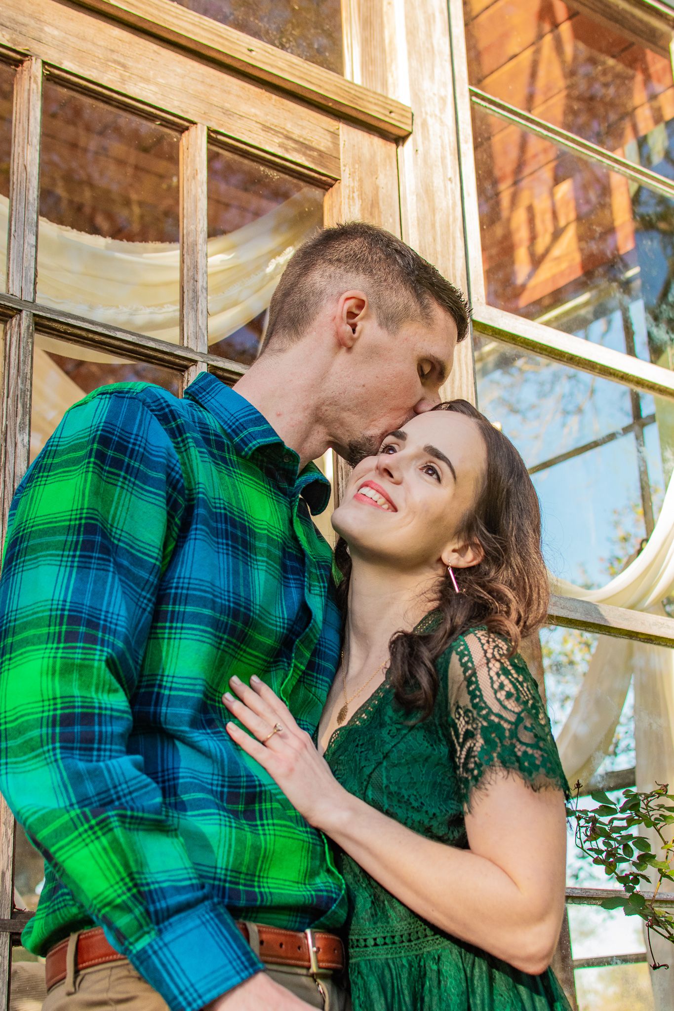 A couple in green clothing shares an intimate moment against rustic windows.
