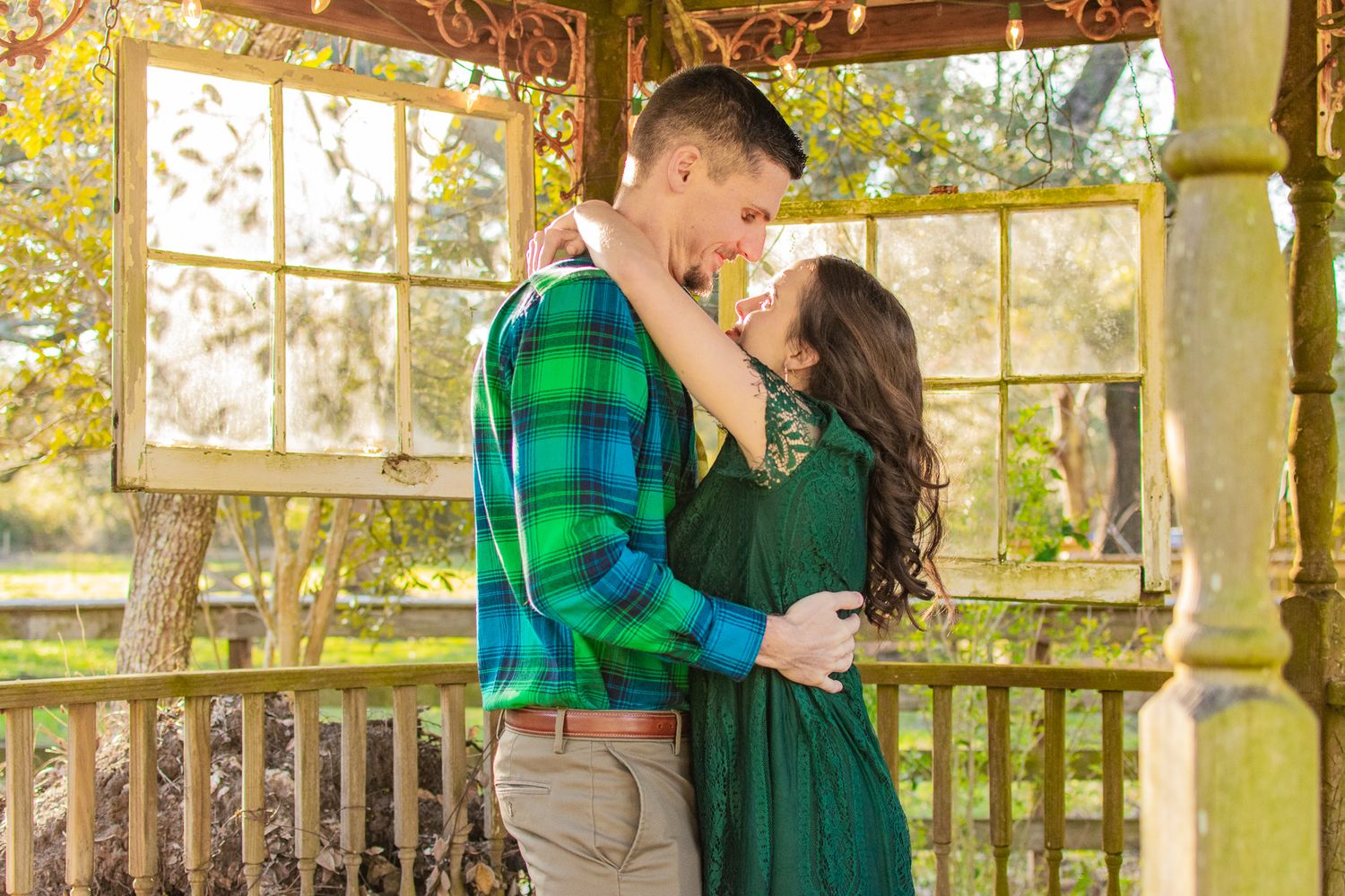 A couple in coordinating green outfits embraces in a wooden gazebo.