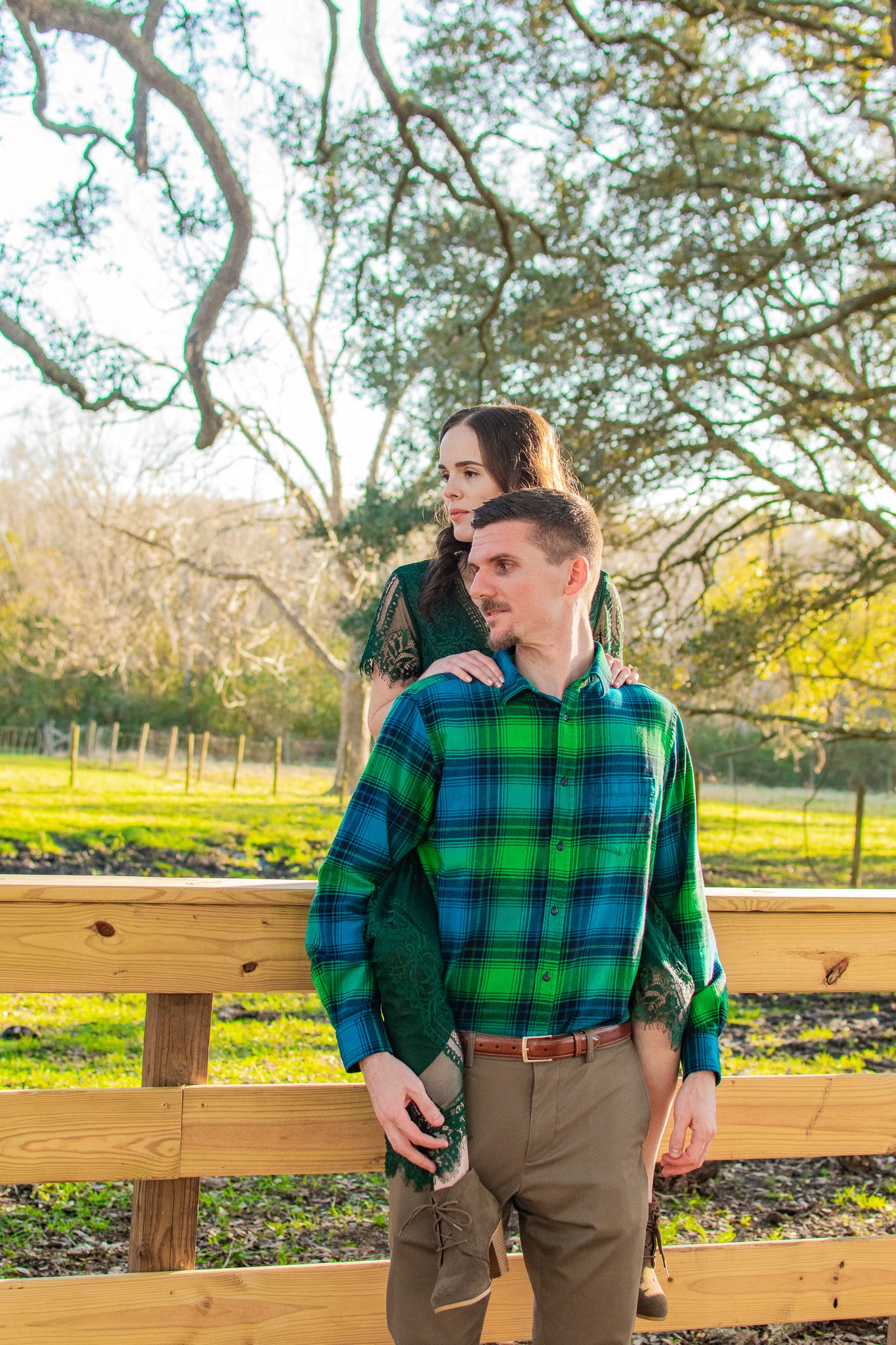Couple in coordinating green outfits pose together near wooden fence in sunny park.