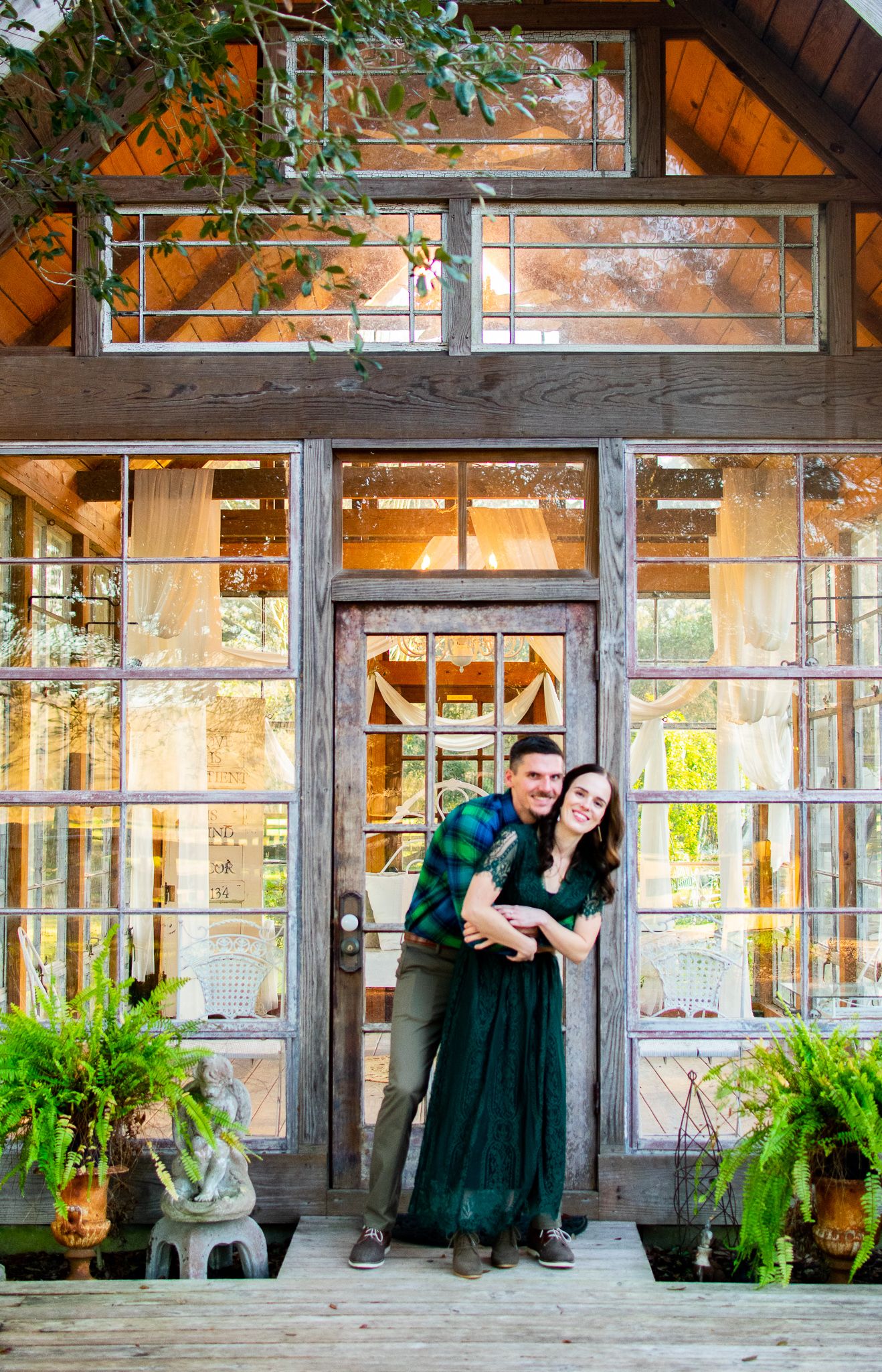 Couple poses in doorway of charming greenhouse with ferns and windows.