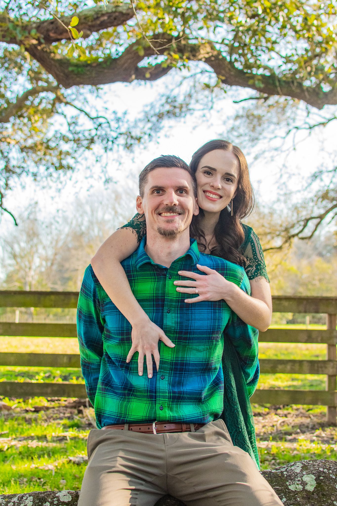 A couple in matching green outfits embraces outdoors near a wooden fence on an autumn day.
