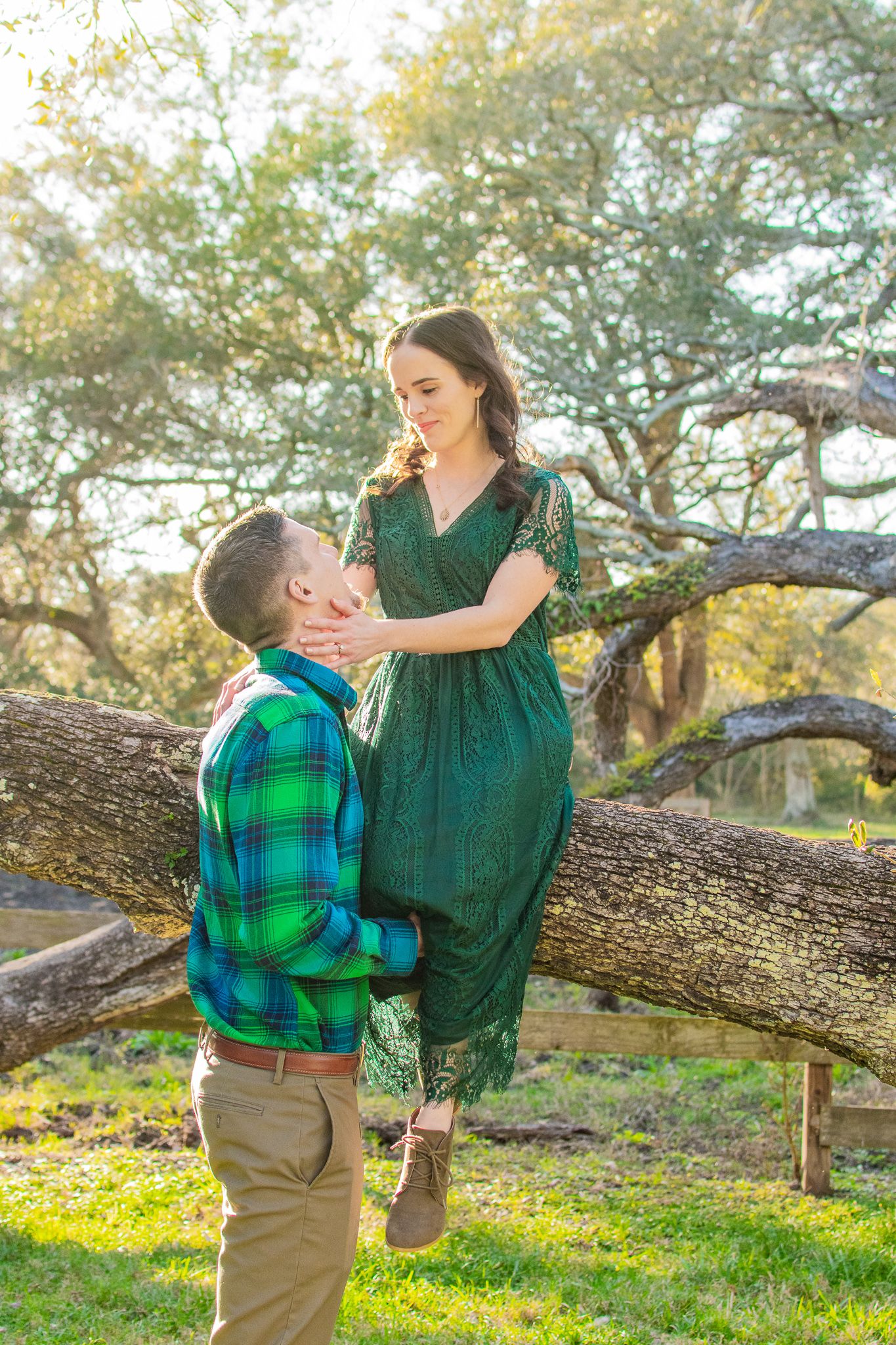 A couple poses romantically by a tree trunk outdoors, with her in a green dress and him in plaid.