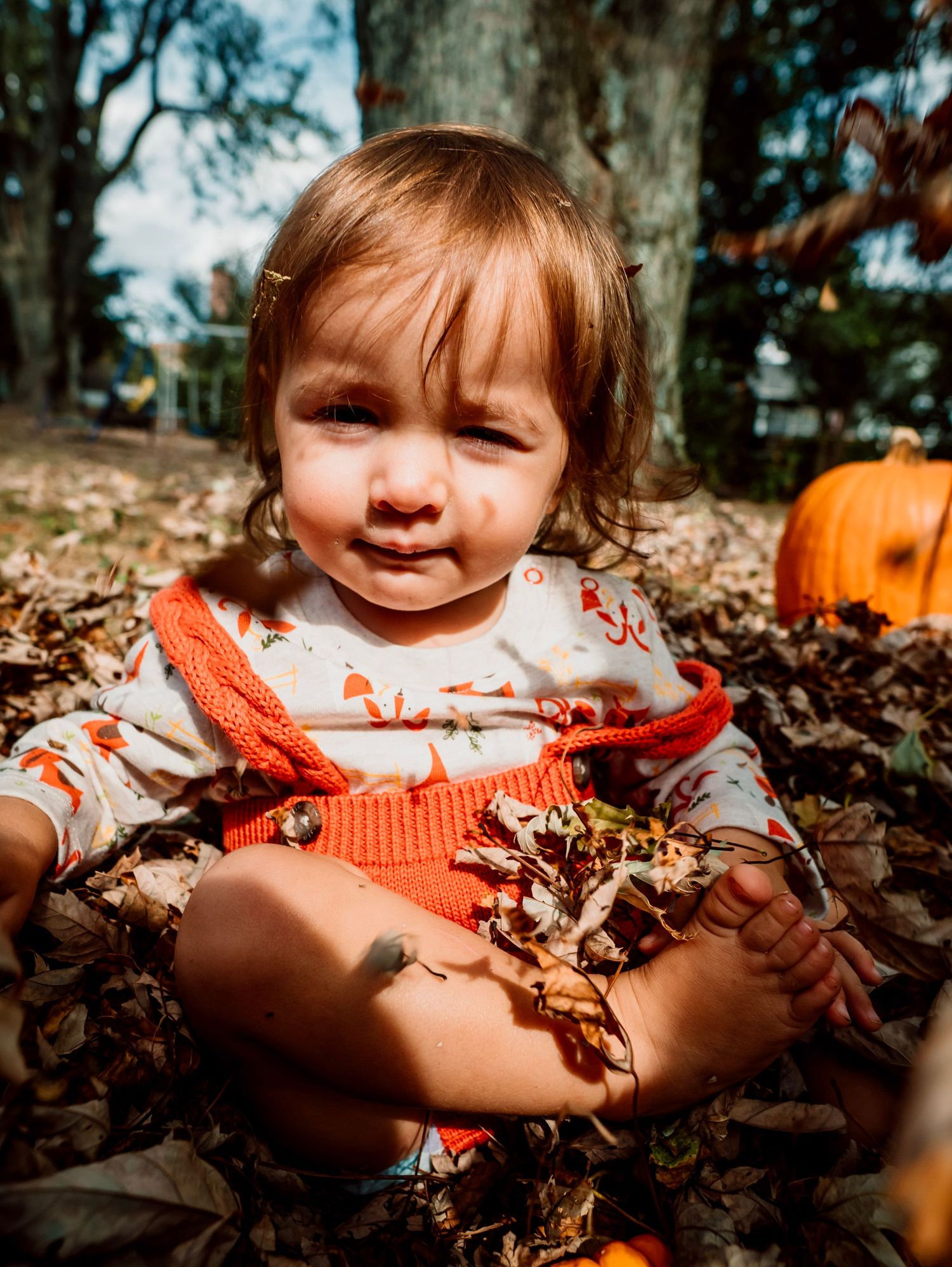 A baby girl plays in the leaves with pumpkins on a beautiful fall day.