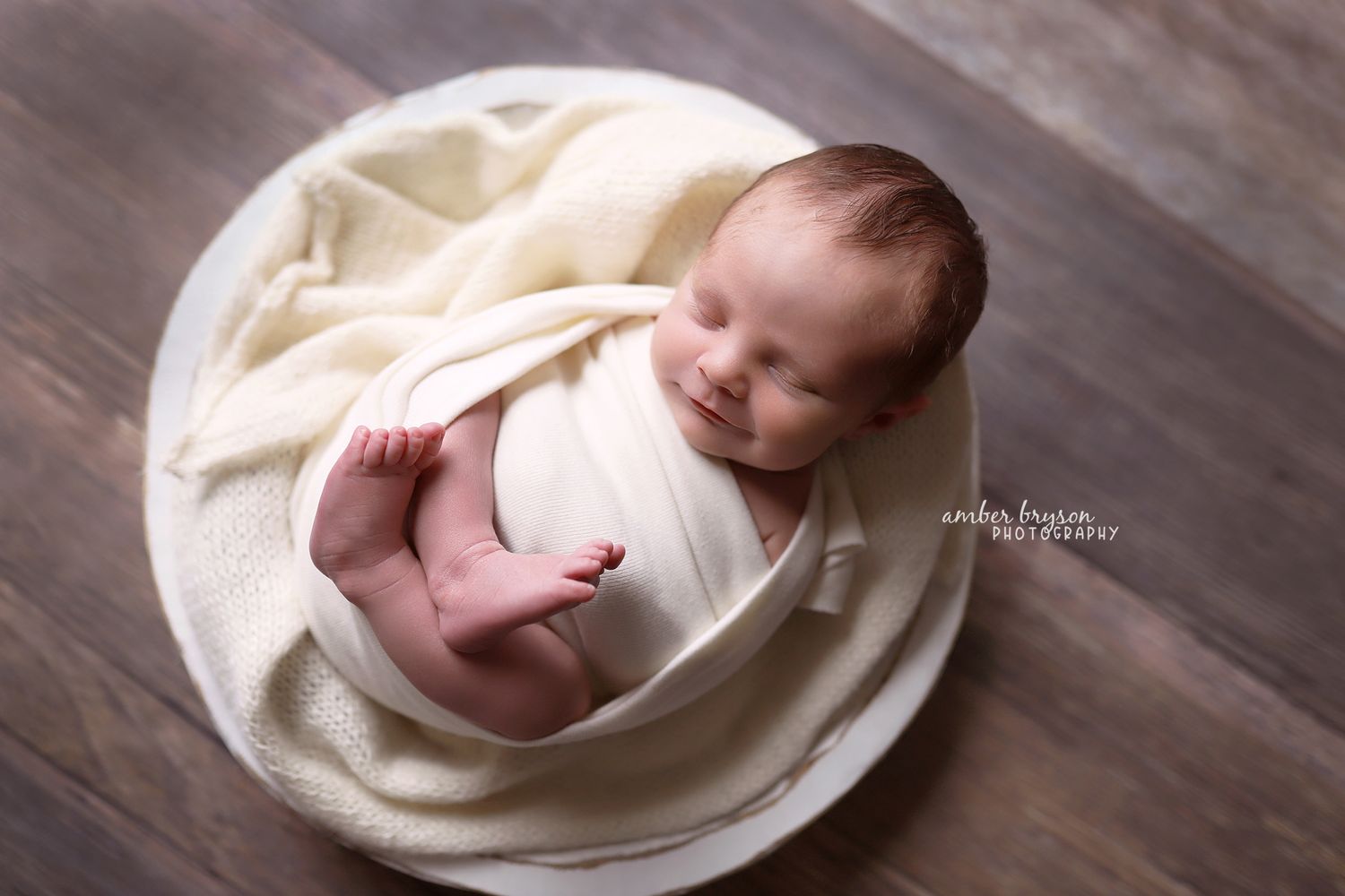 Newborn baby wrapped in cream blanket lying in white fabric nest on wooden floor.