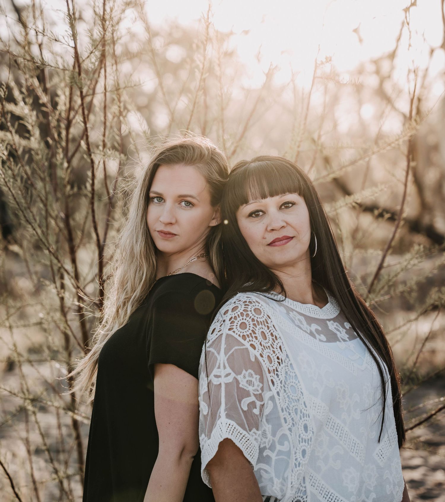 Two women in contrasting black and white outfits pose in sunlit natural setting.