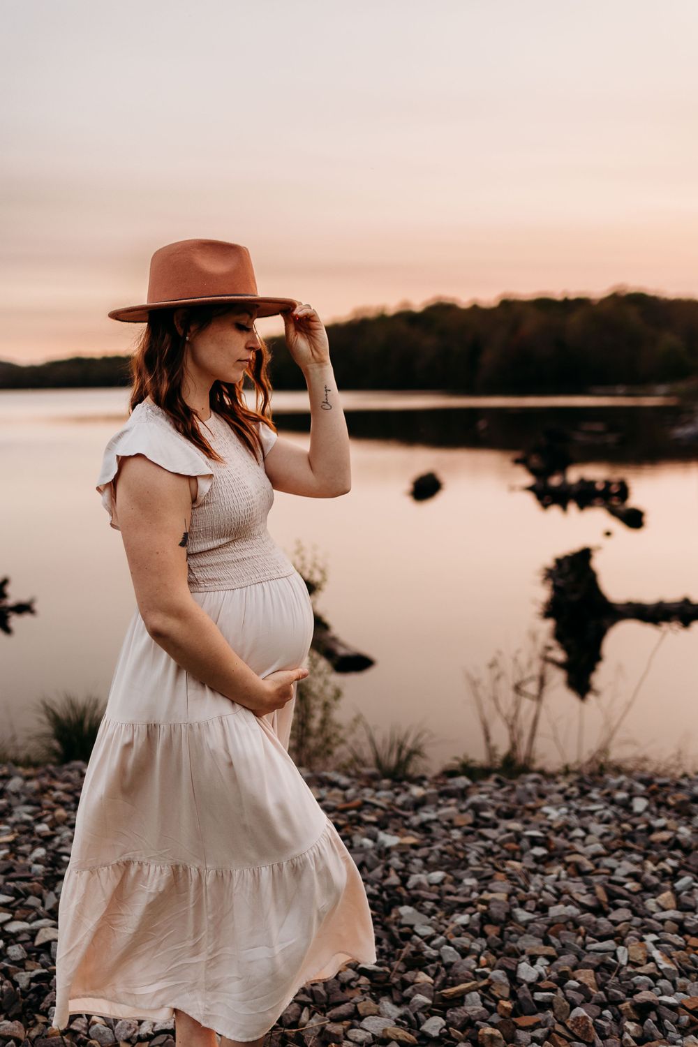 Expectant mother in flowy dress and hat poses by lake at sunset.