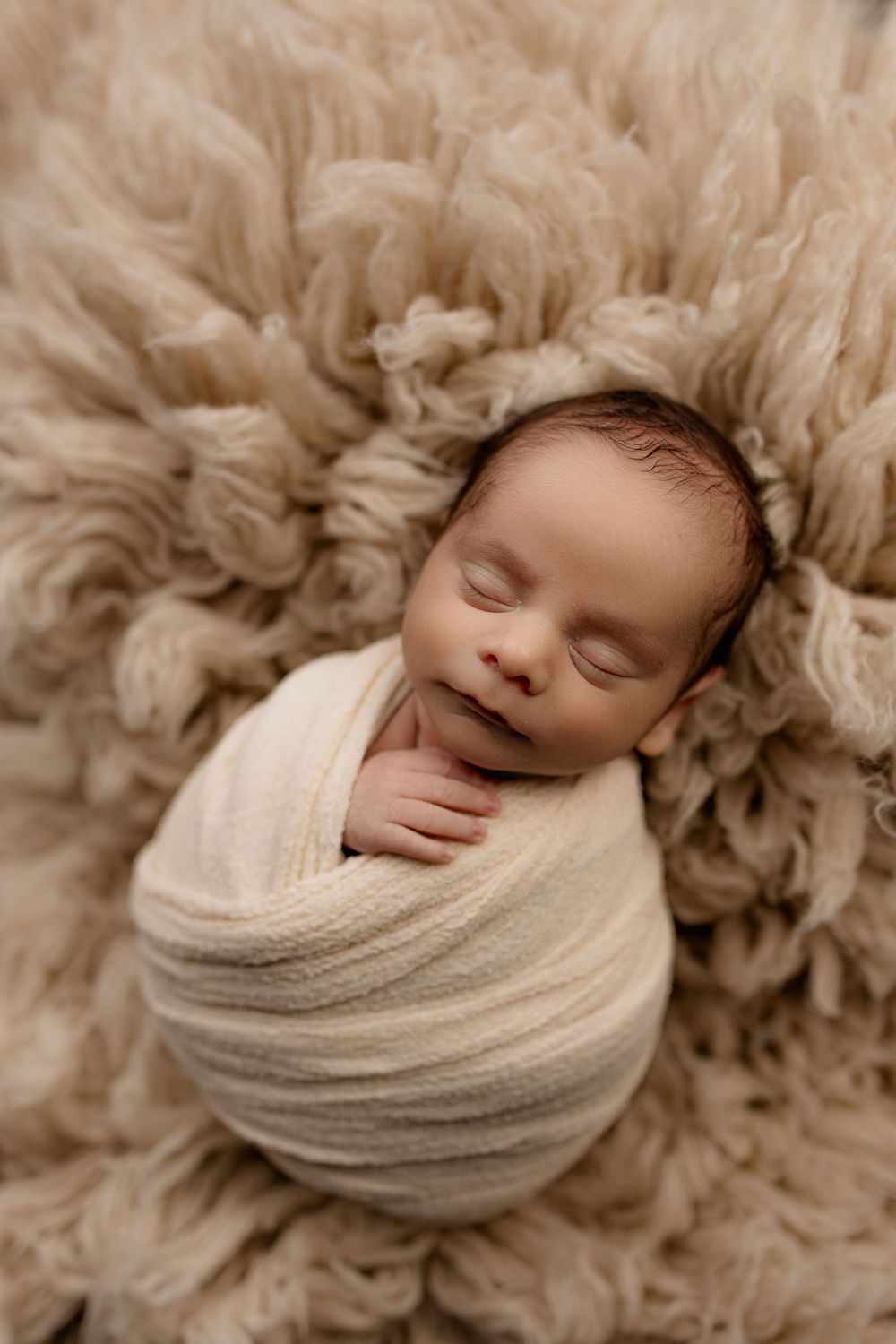 Sleeping newborn wrapped in cream colored swaddle on textured furry backdrop.