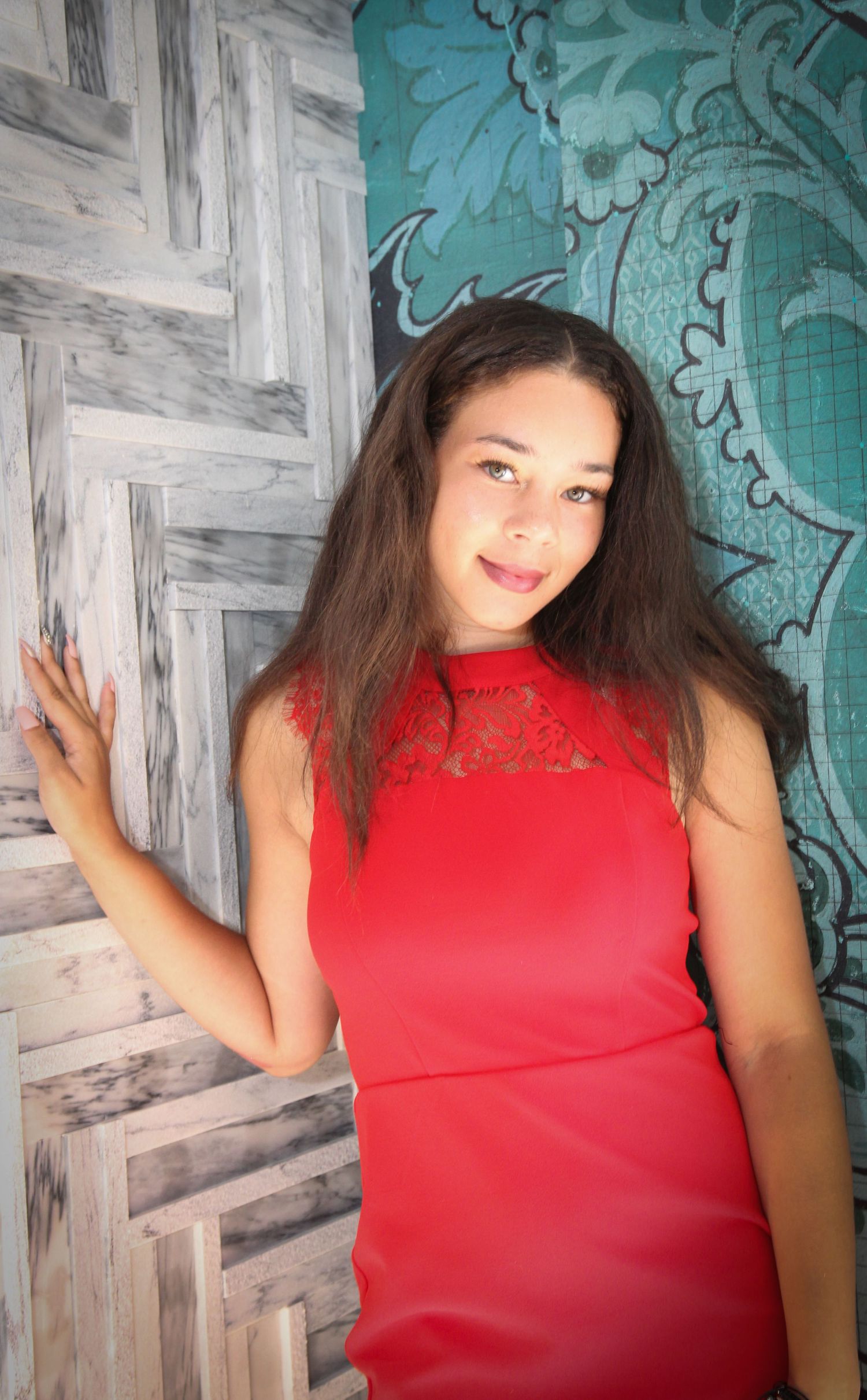 A woman in a red lace dress poses against a turquoise and gray patterned wall.