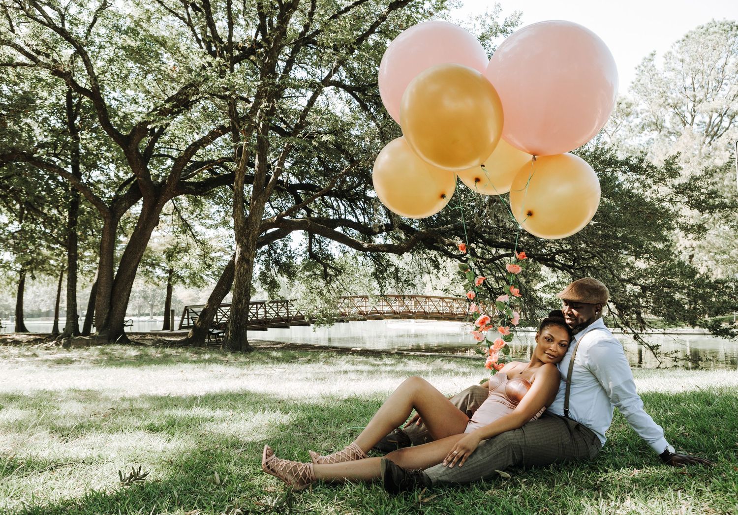 A couple relaxes under a tree in a sunny park with pink and yellow balloons floating above them.