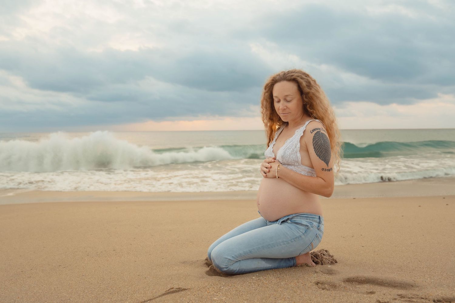 Expectant mother in jeans kneeling on beach at golden hour.