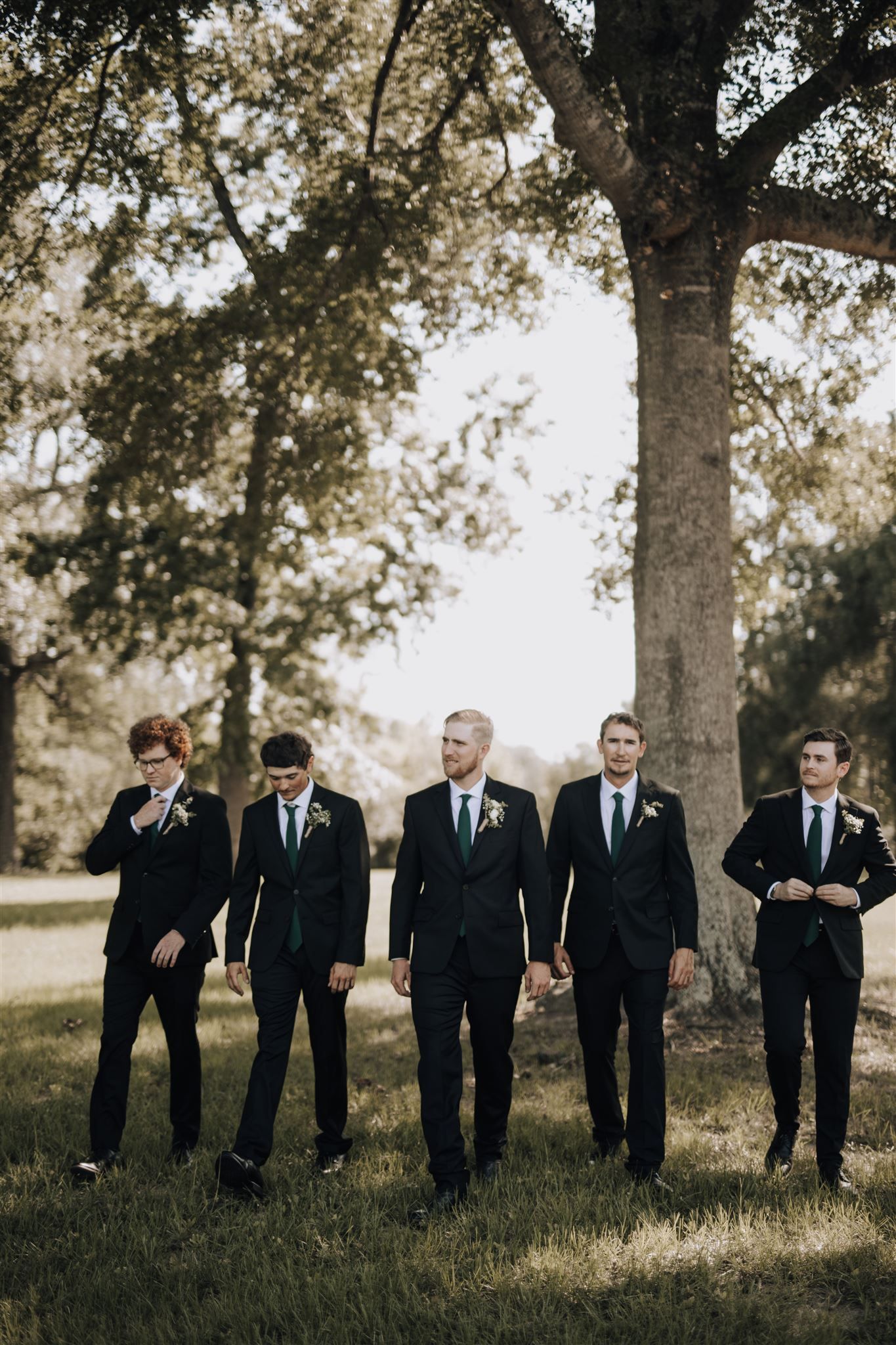 Groomsmen in dark suits stand together under large tree in outdoor setting.