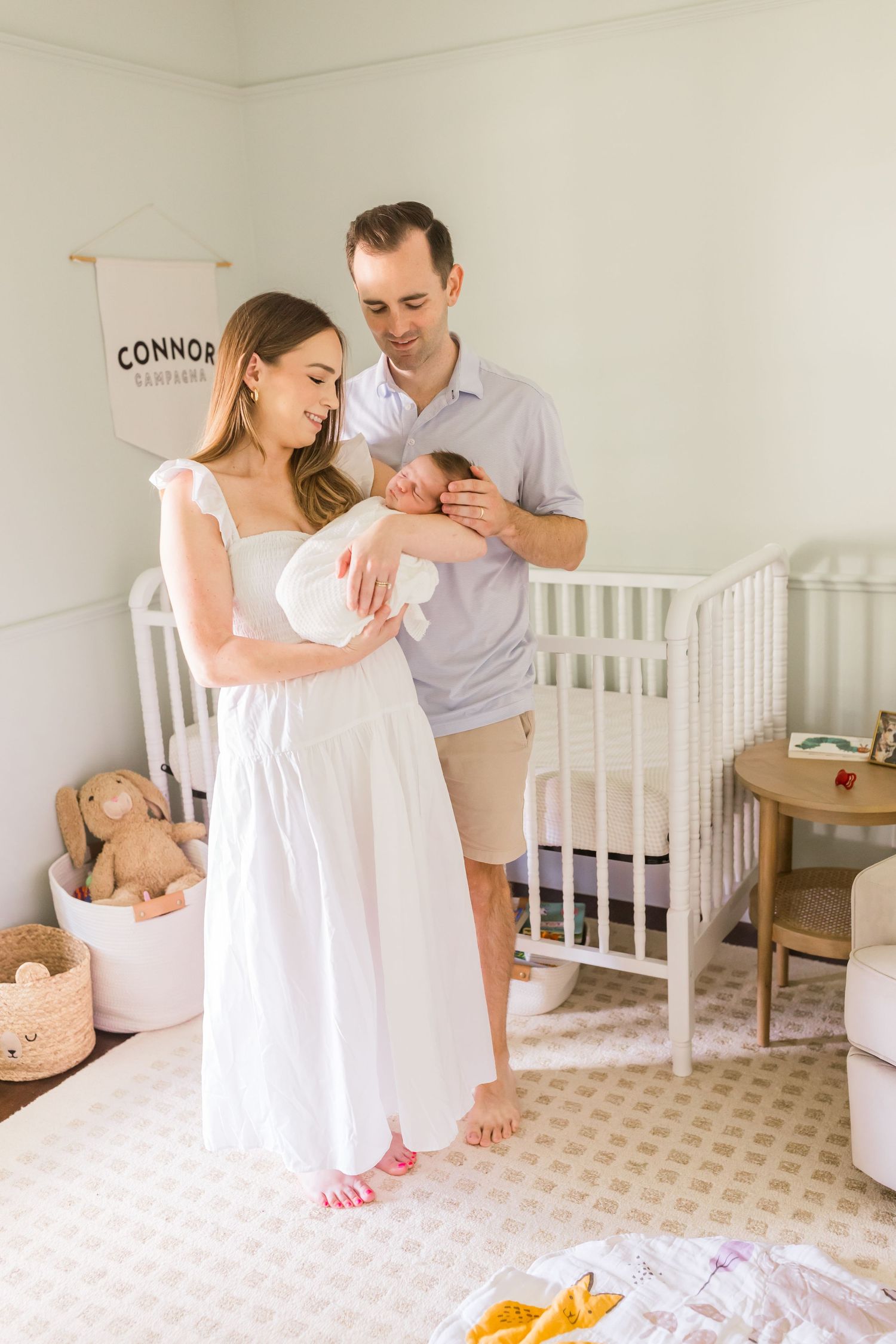 Parents holding newborn baby in white nursery with crib and decorative details.