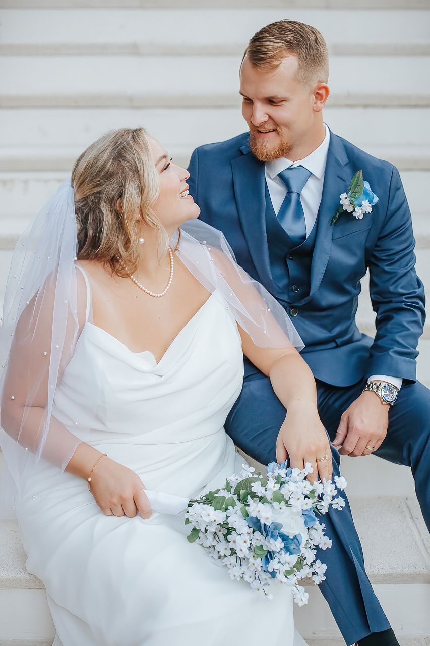Wedding couple poses together with blue and white floral bouquet against light backdrop.