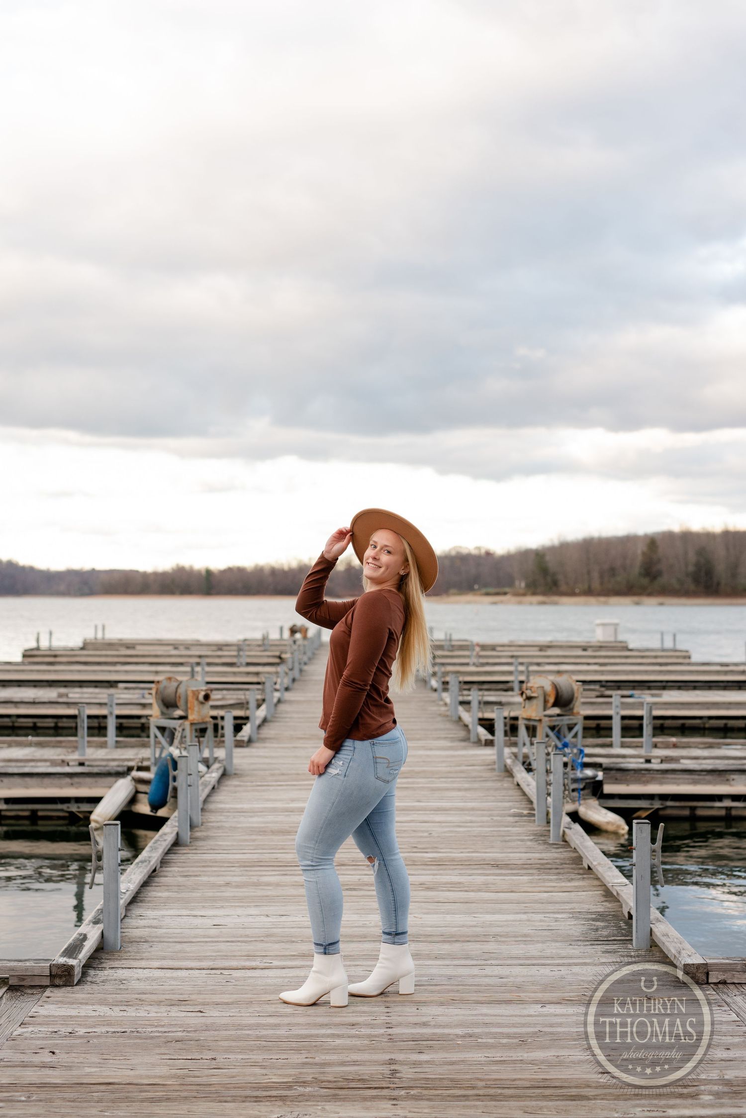 Person in brown sweater and hat posing on wooden dock by lake during sunset.