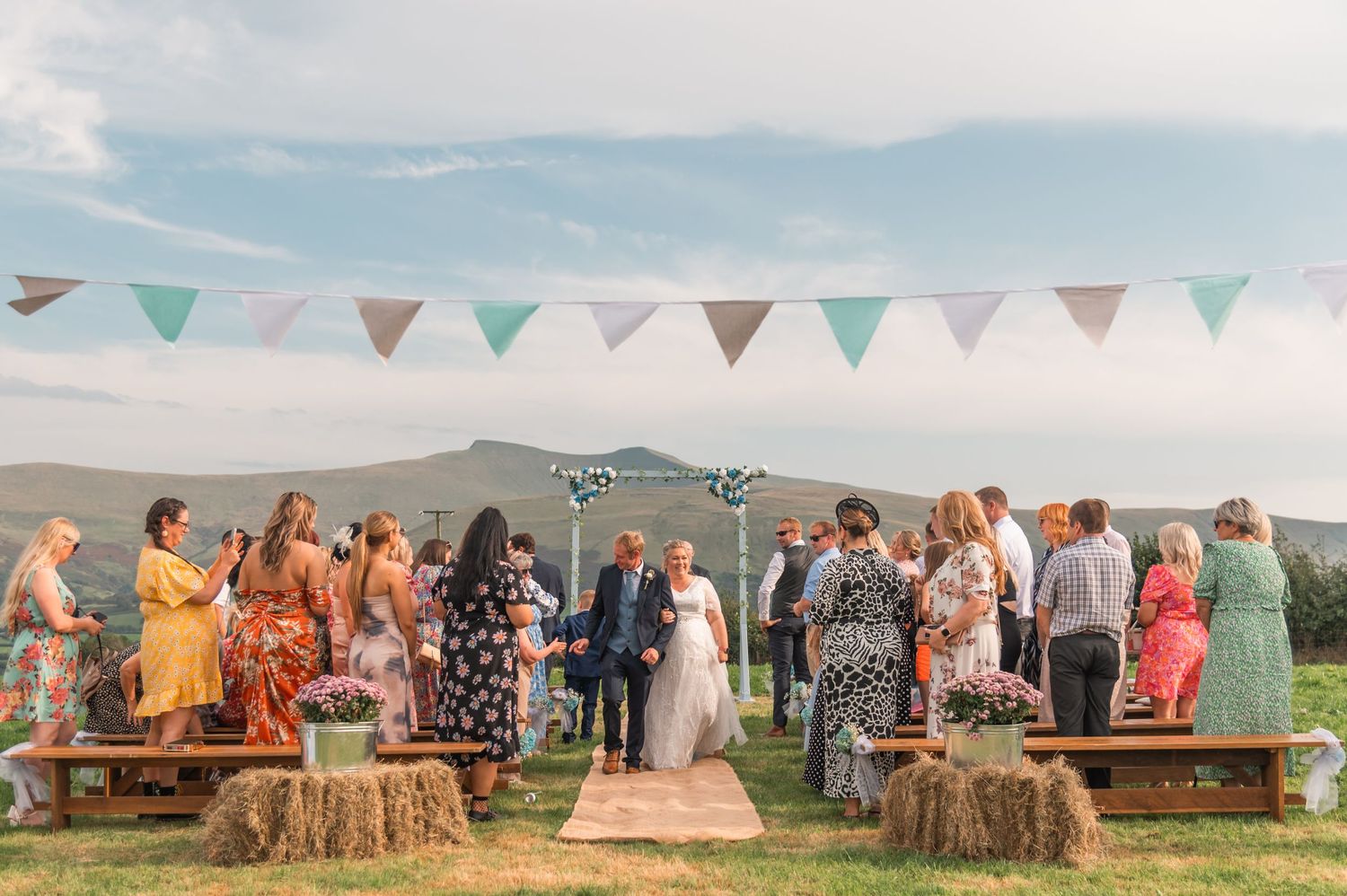 Wedding guests seated on hay bales at an outdoor ceremony with mountain views and festive bunting decorations.