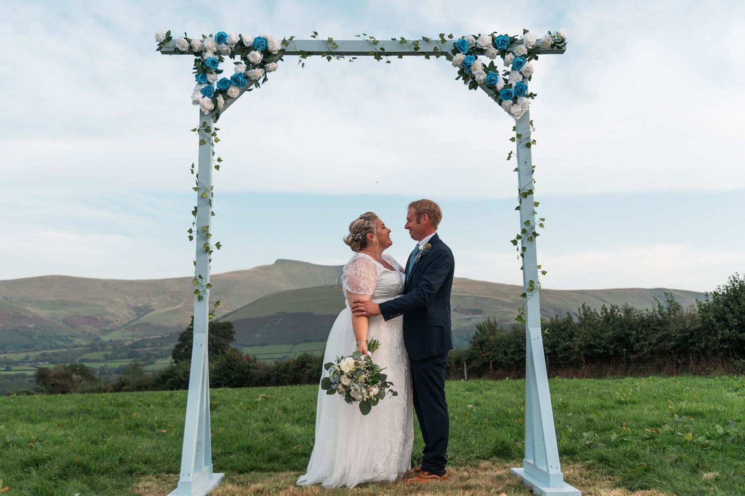 Outdoor wedding ceremony under a floral arch with white fabric draping and mountain backdrop.