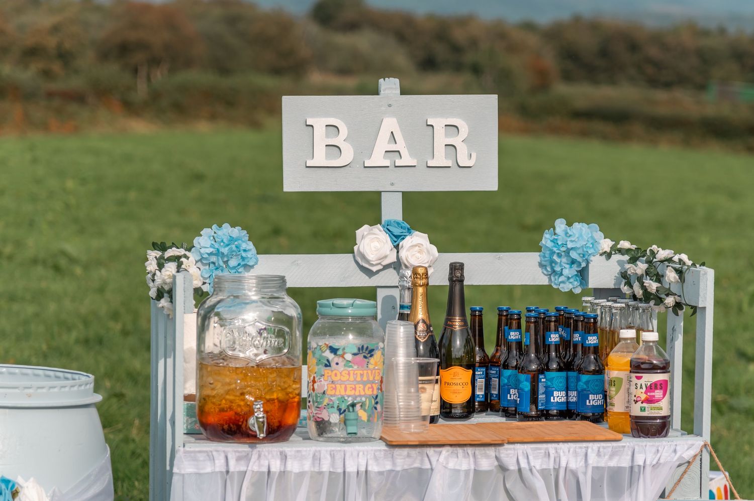 Wedding bar setup with drink dispensers and blue floral decorations at outdoor reception.