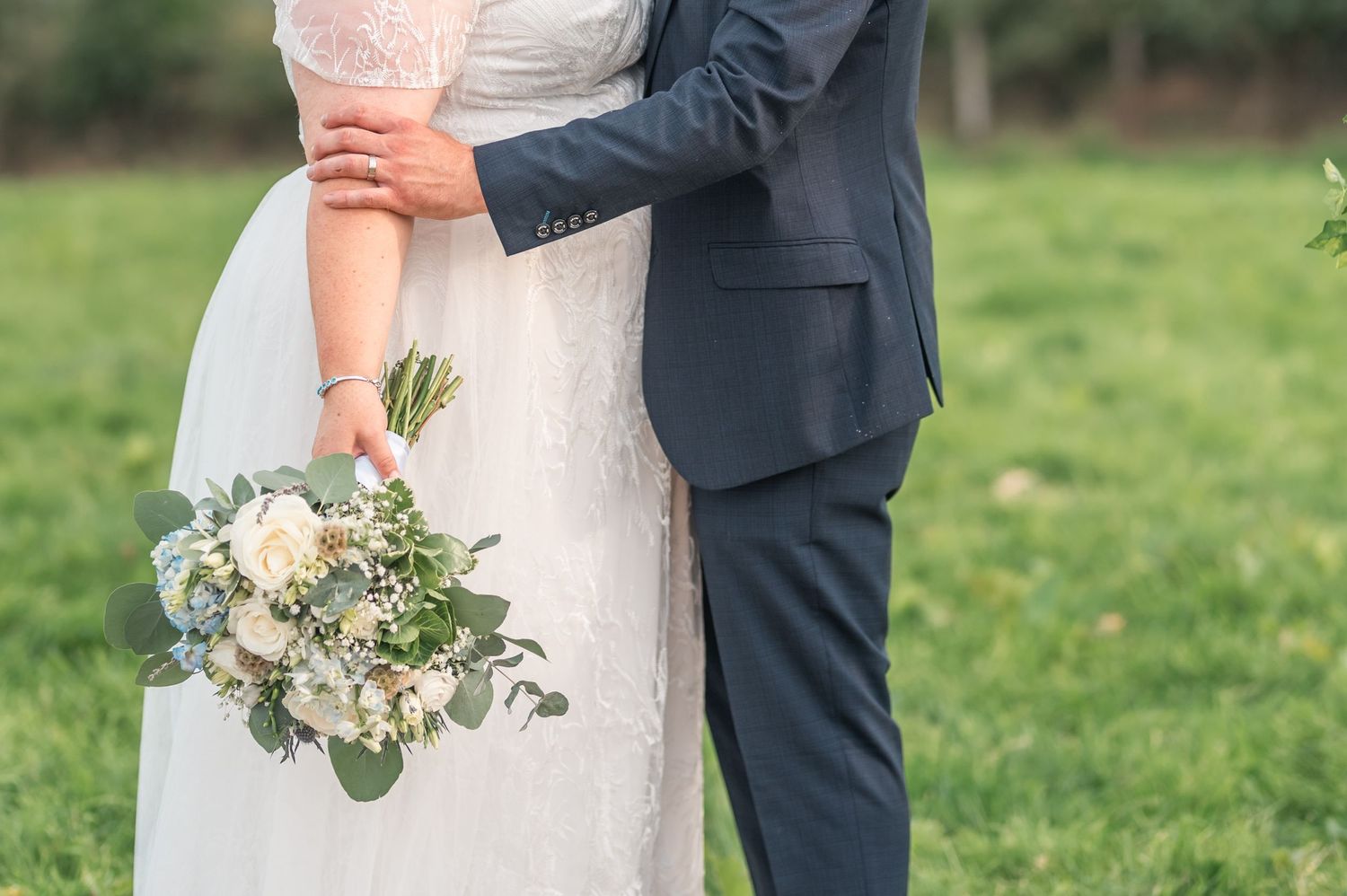A bride in white gown and groom in navy suit embrace outdoors on their wedding day.