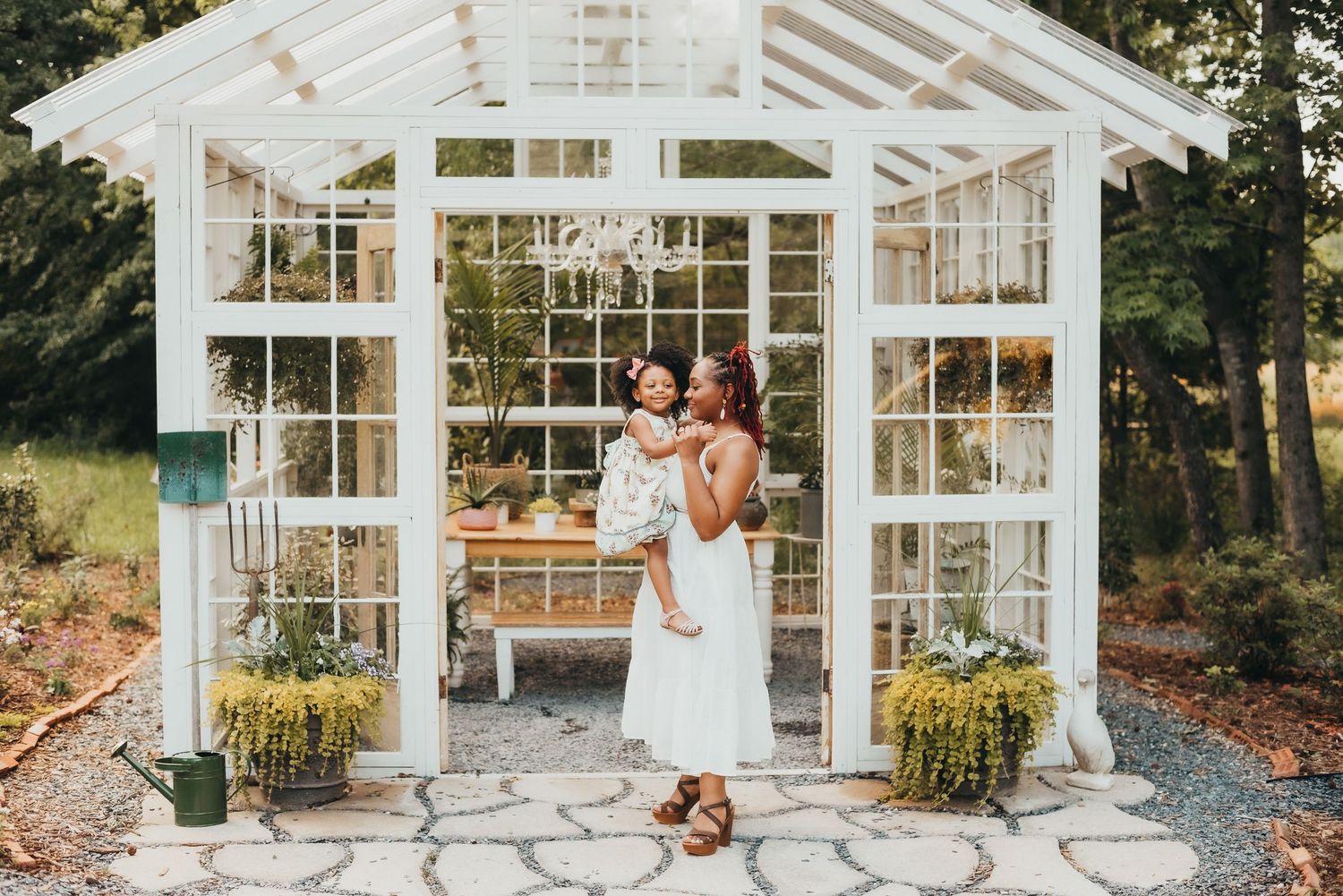 Mother and daughter in white dresses pose outside a charming white greenhouse garden structure.