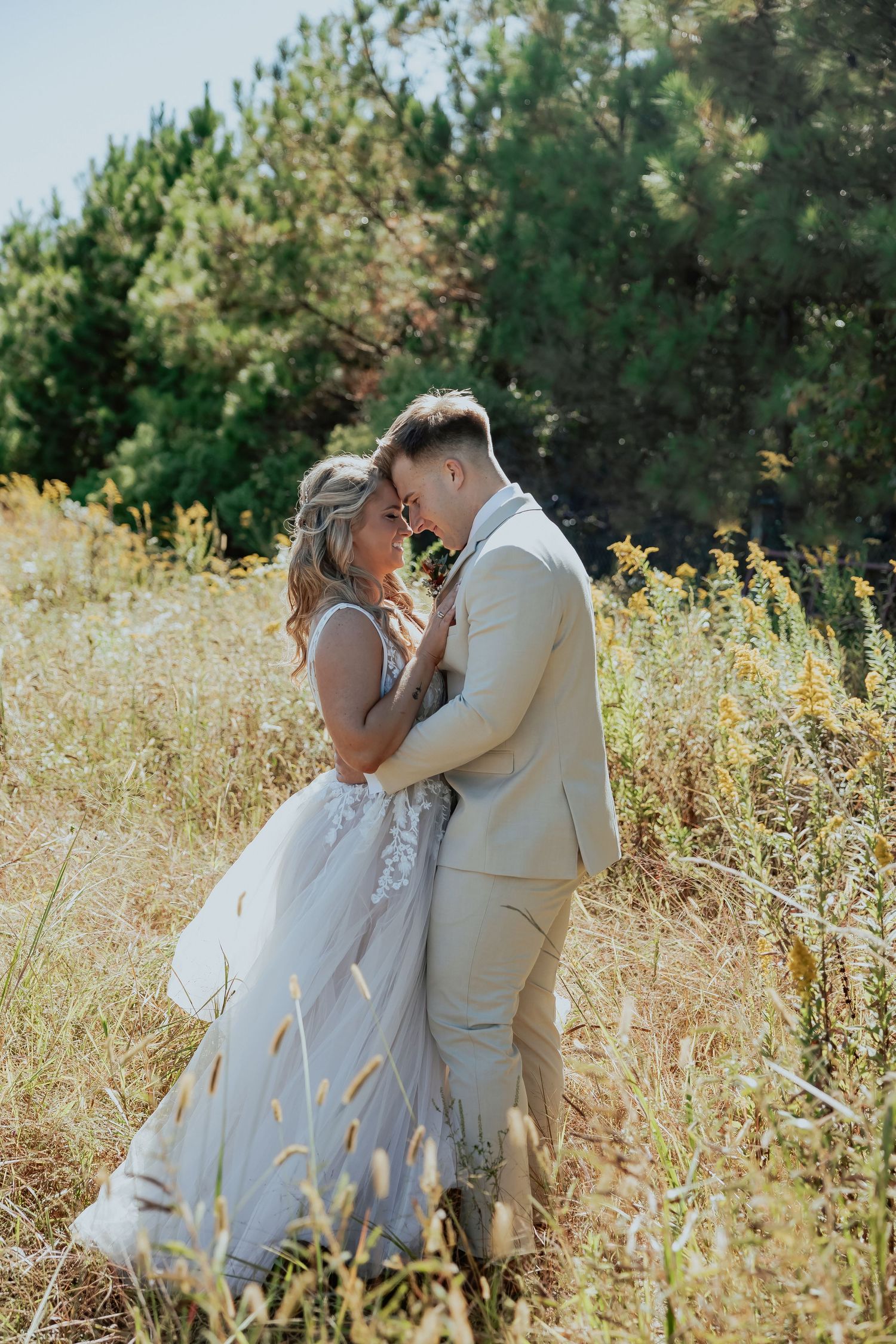 A romantic outdoor photoshoot in a sunlit field with tall grass and pine trees in the background.
