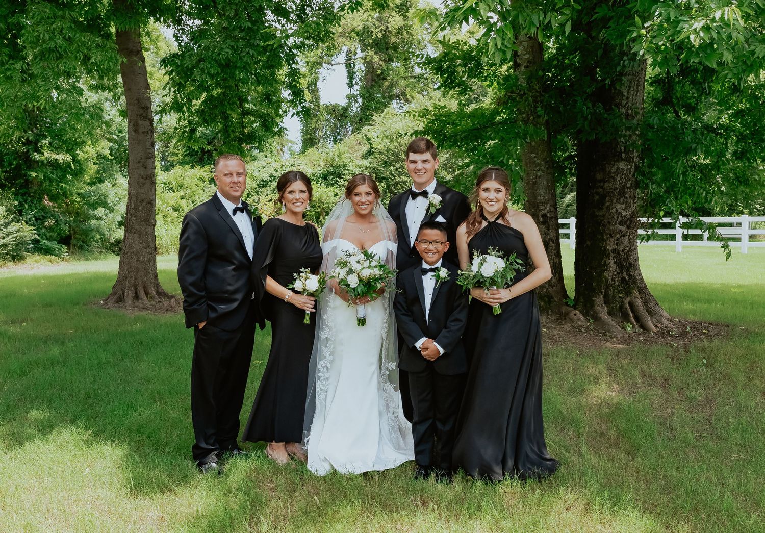 A wedding party poses together under green trees for formal group photos.
