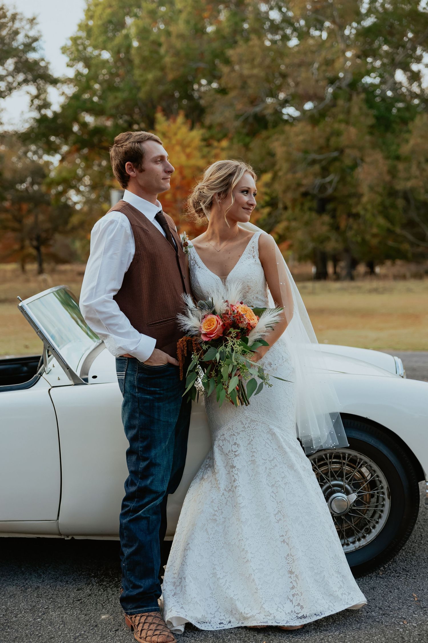 A wedding couple poses beside a classic vintage car with the bride holding a vibrant autumn bouquet.