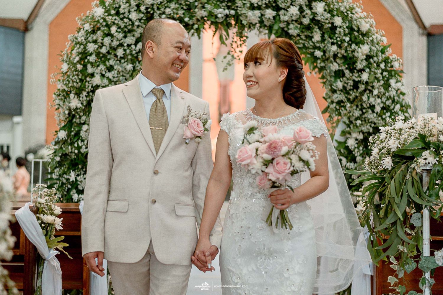 Wedding couple sharing joyful moment under floral arch with white flowers.